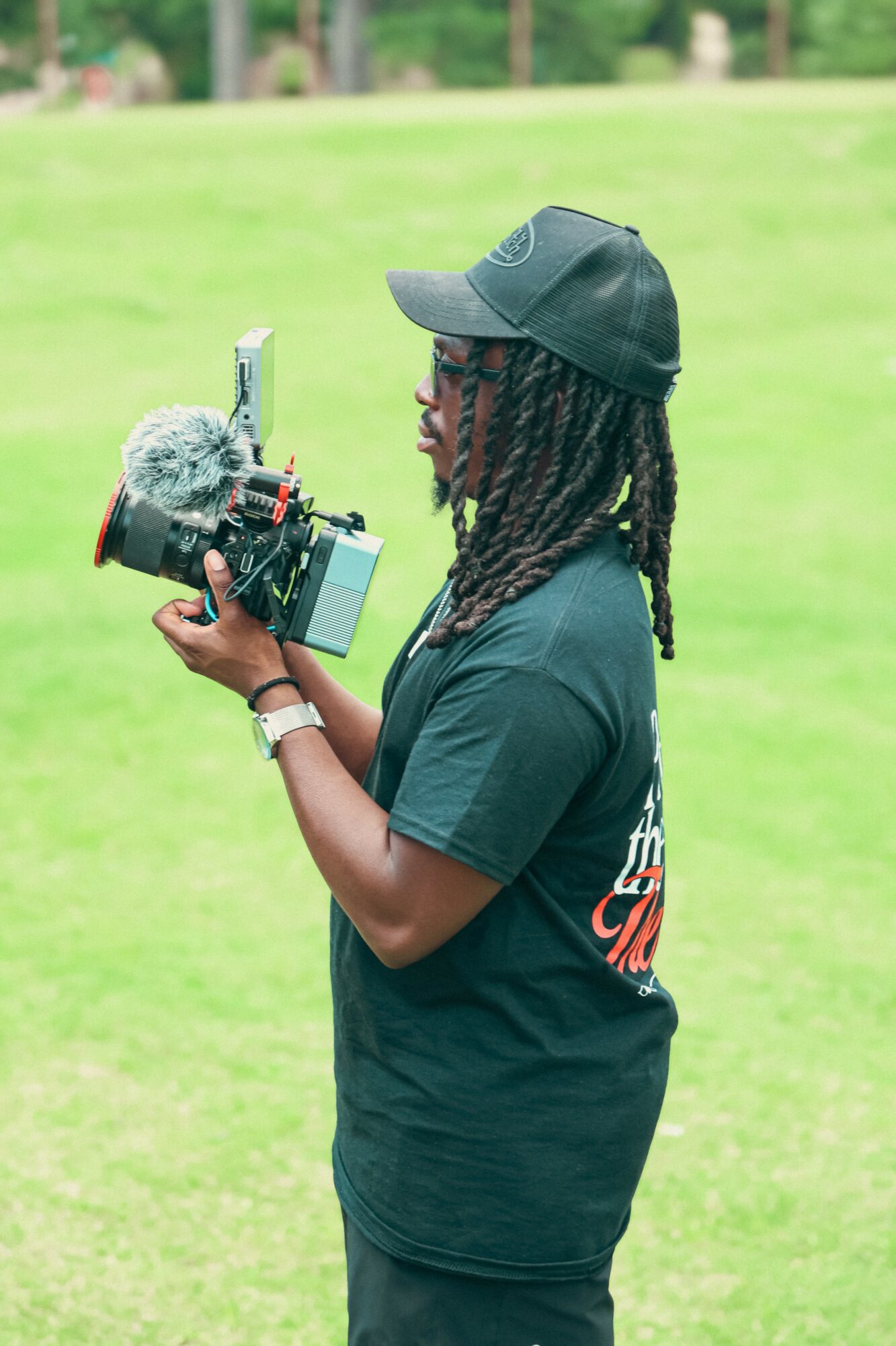 Person with dreadlocks wearing a black cap and dark t-shirt holding a camera with a microphone outdoors on green grass