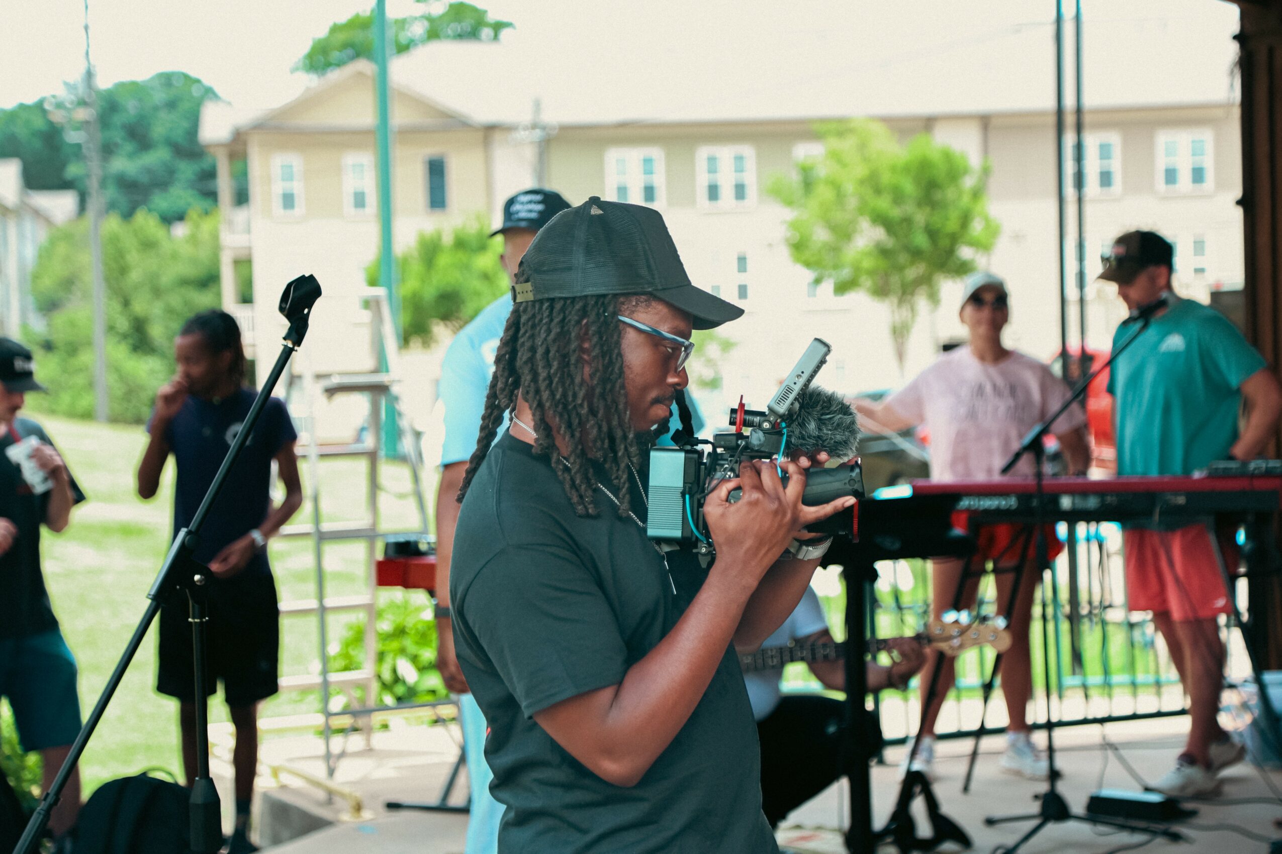 Person with long braided hair and glasses operating a camera at an outdoor event, with people and musical equipment in background.