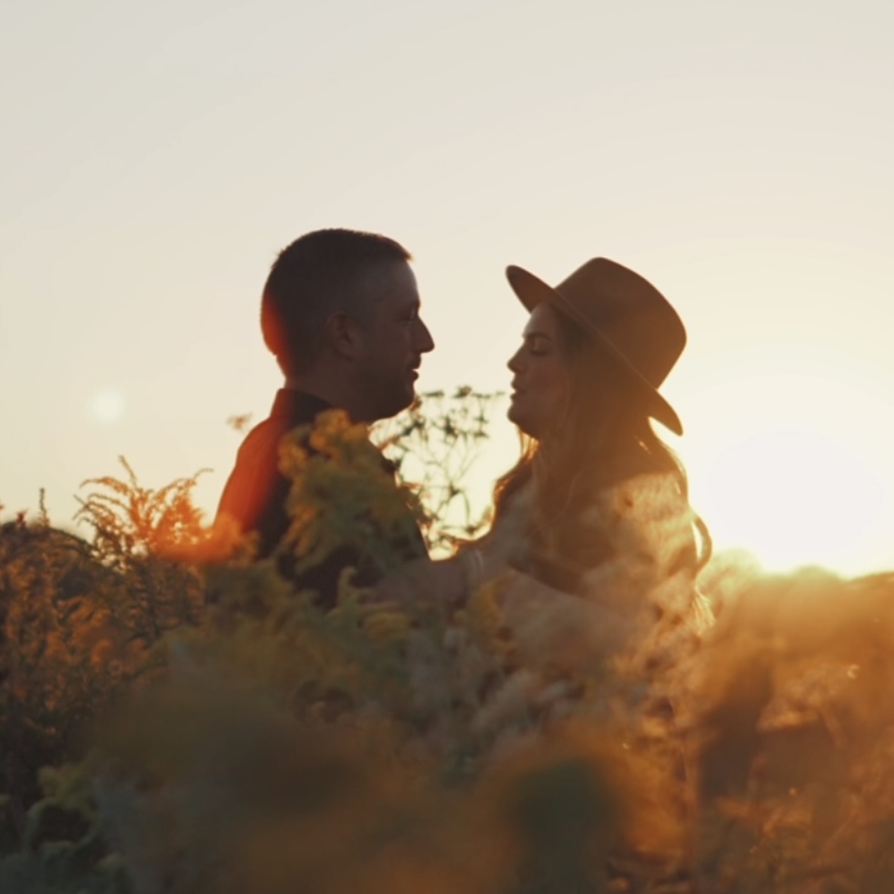 Silhouettes of a man and woman facing each other outdoors during sunset, surrounded by plants and flowers.