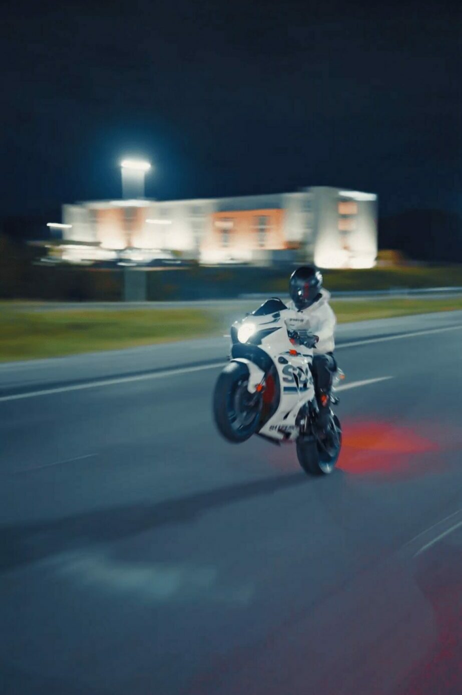 Motorcycle rider in motion on a road at night with a building and bright light in background.