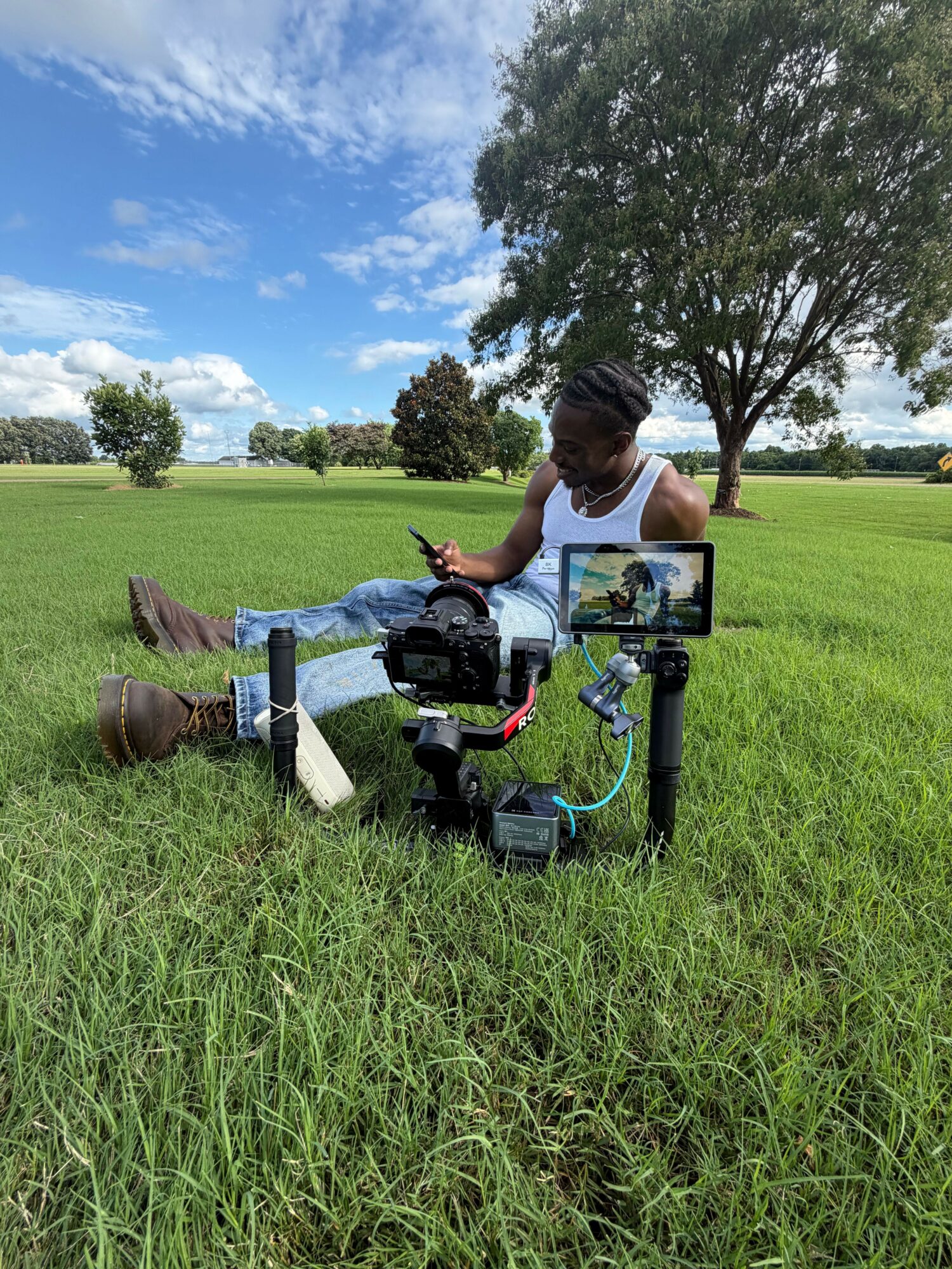 Person operating a camera on a grassy field with trees and blue sky in background.