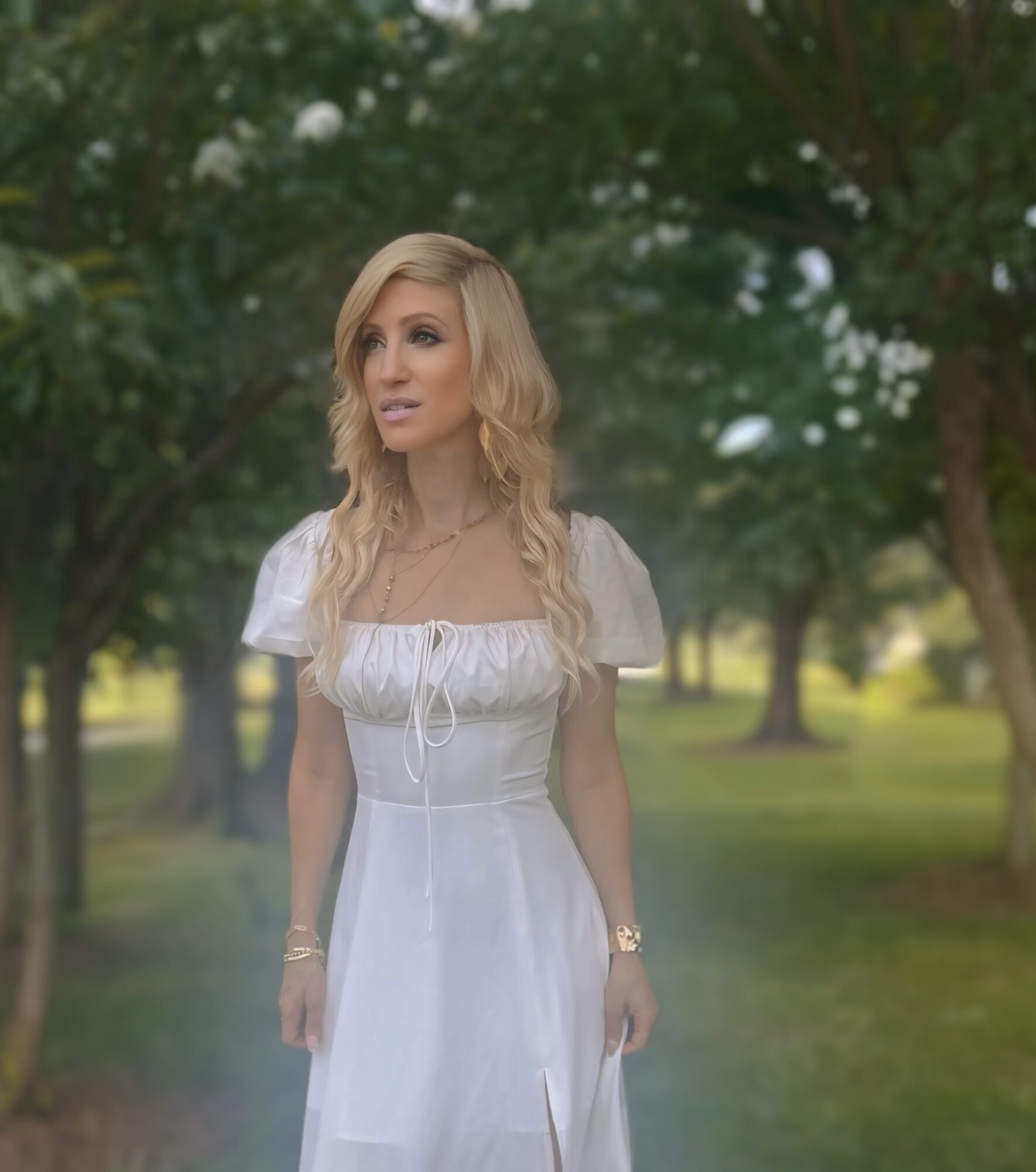 Woman in white dress standing outdoors with trees in background, looking to the side.