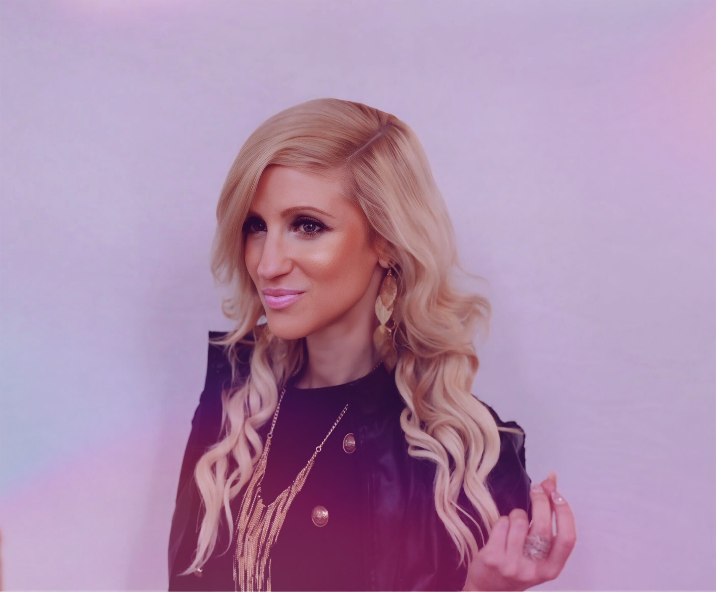 Woman with long blonde hair and earrings, wearing a dark jacket, smiling, against a light background.