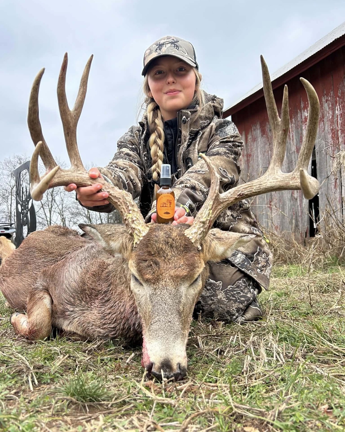 Child in camouflage clothing kneels behind a large deer with antlers, outdoors near a red building, holding the antlers.
