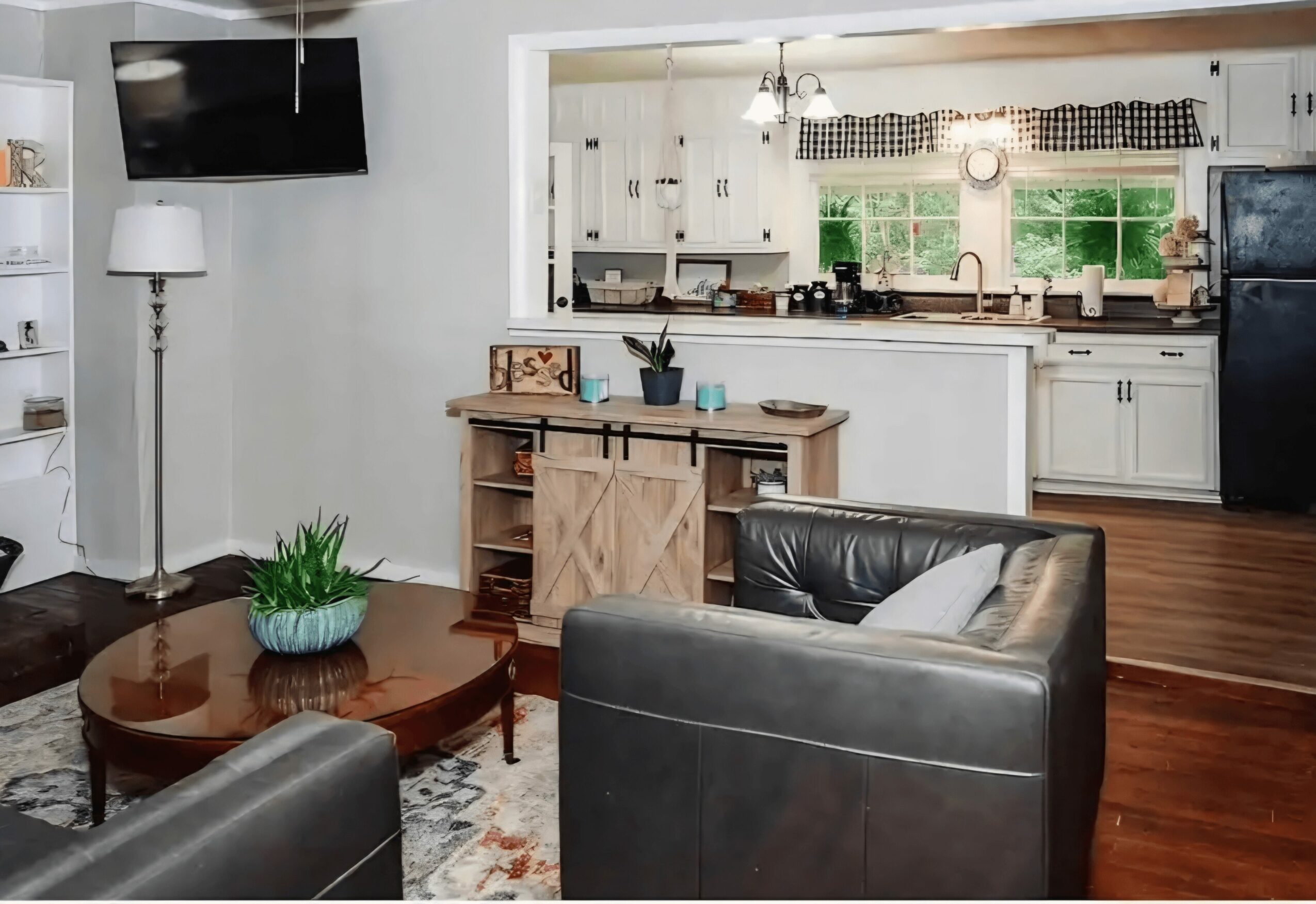 Living room with black armchairs, wooden coffee table, potted plant, and a view into the kitchen with white cabinets and windows.