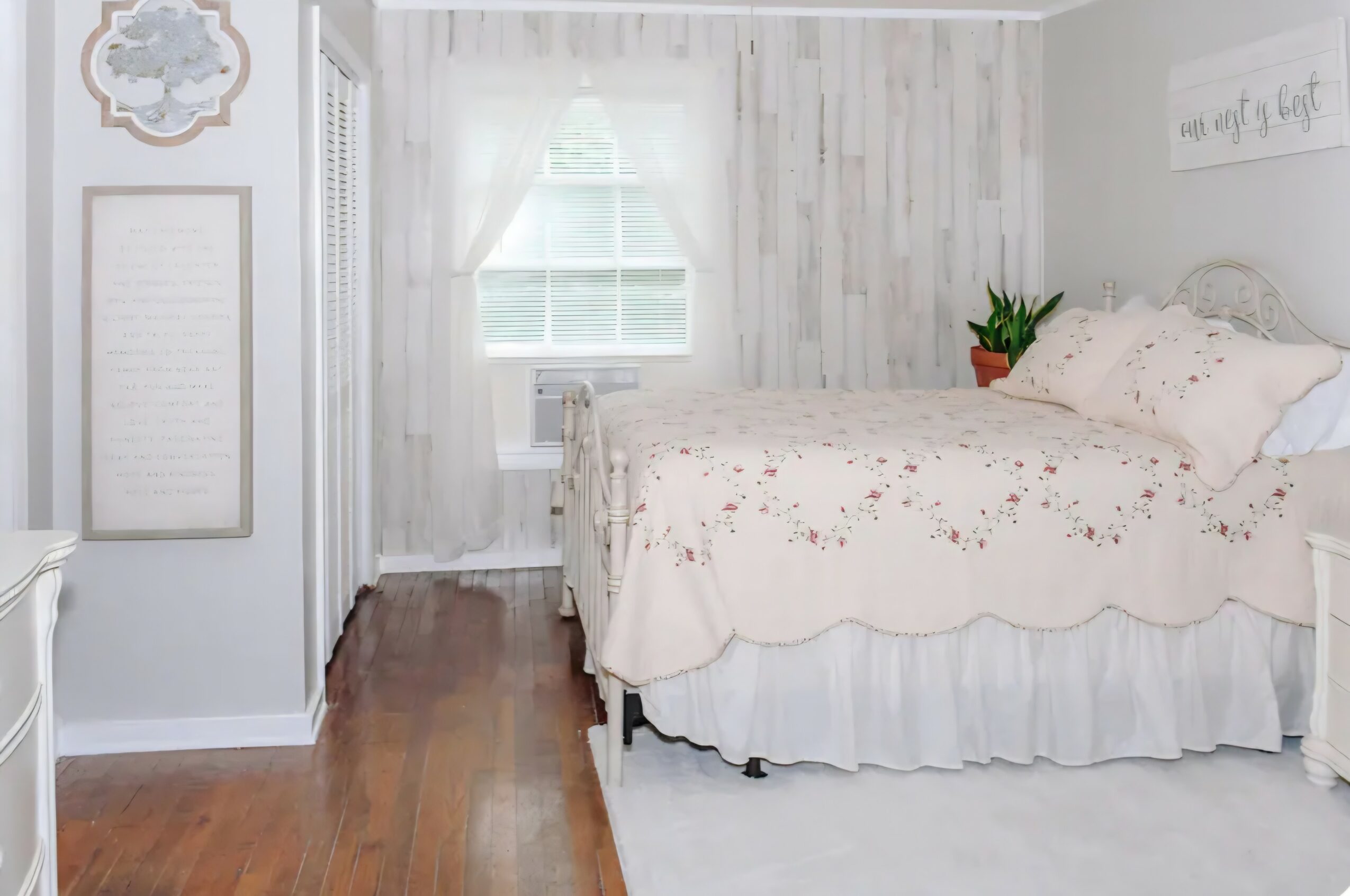 Bedroom with white walls, window with curtains, bed with floral bedding, and wooden floor.