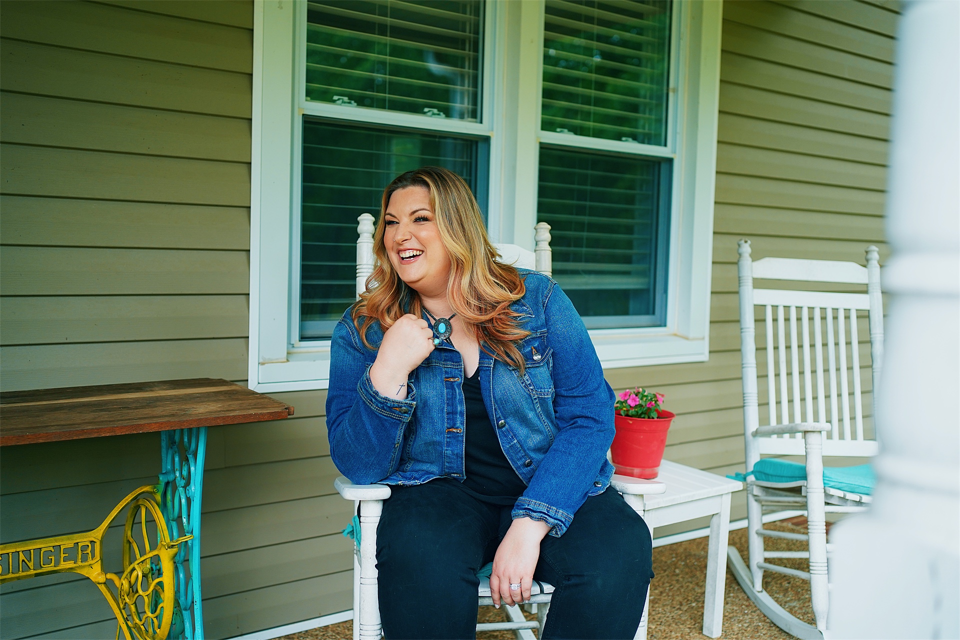 Woman with long hair smiling, sitting outdoors near a house with green siding and white window frames, wearing a denim jacket.