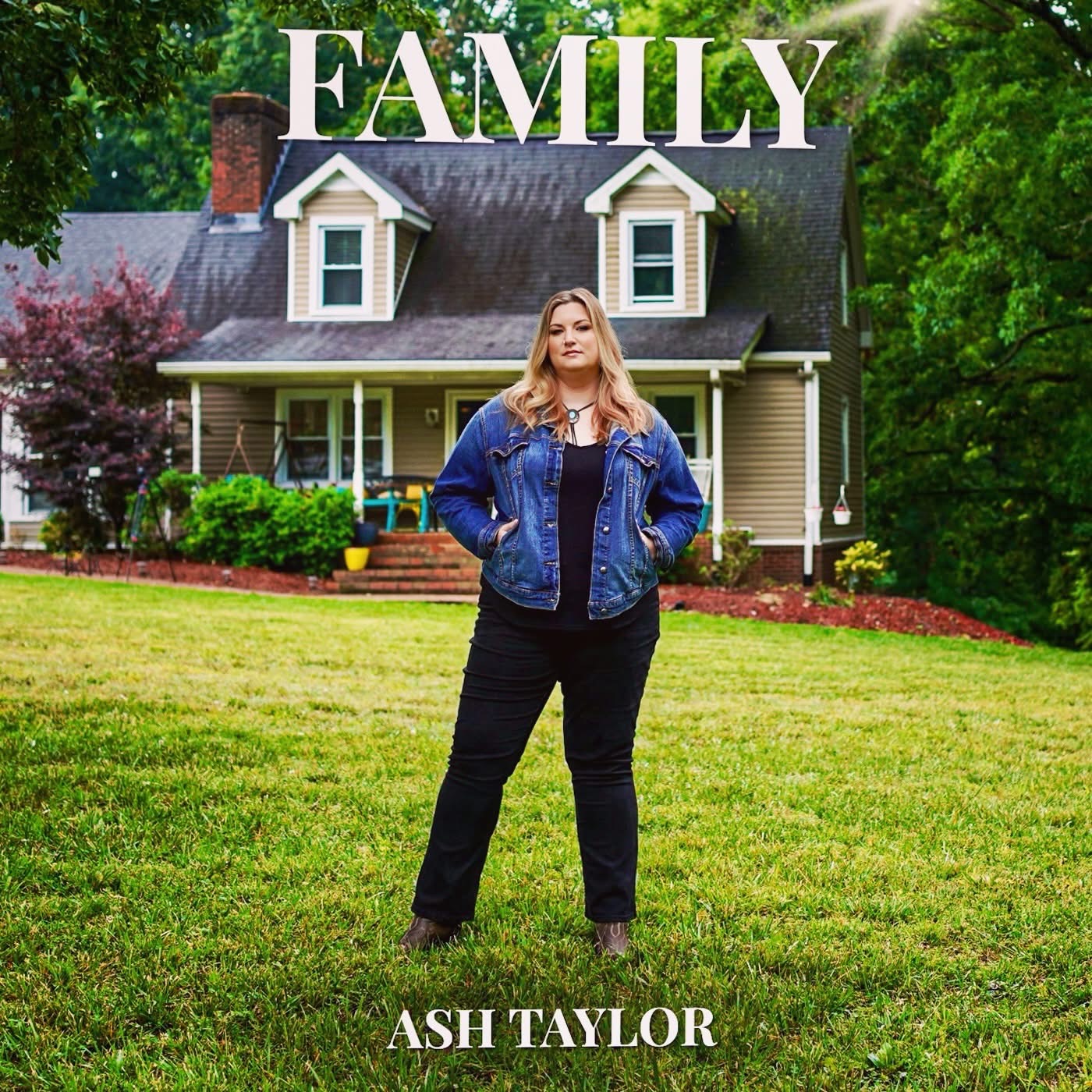 Woman standing on lawn in front of house with large family sign above, trees in background, wearing denim jacket and black pants.