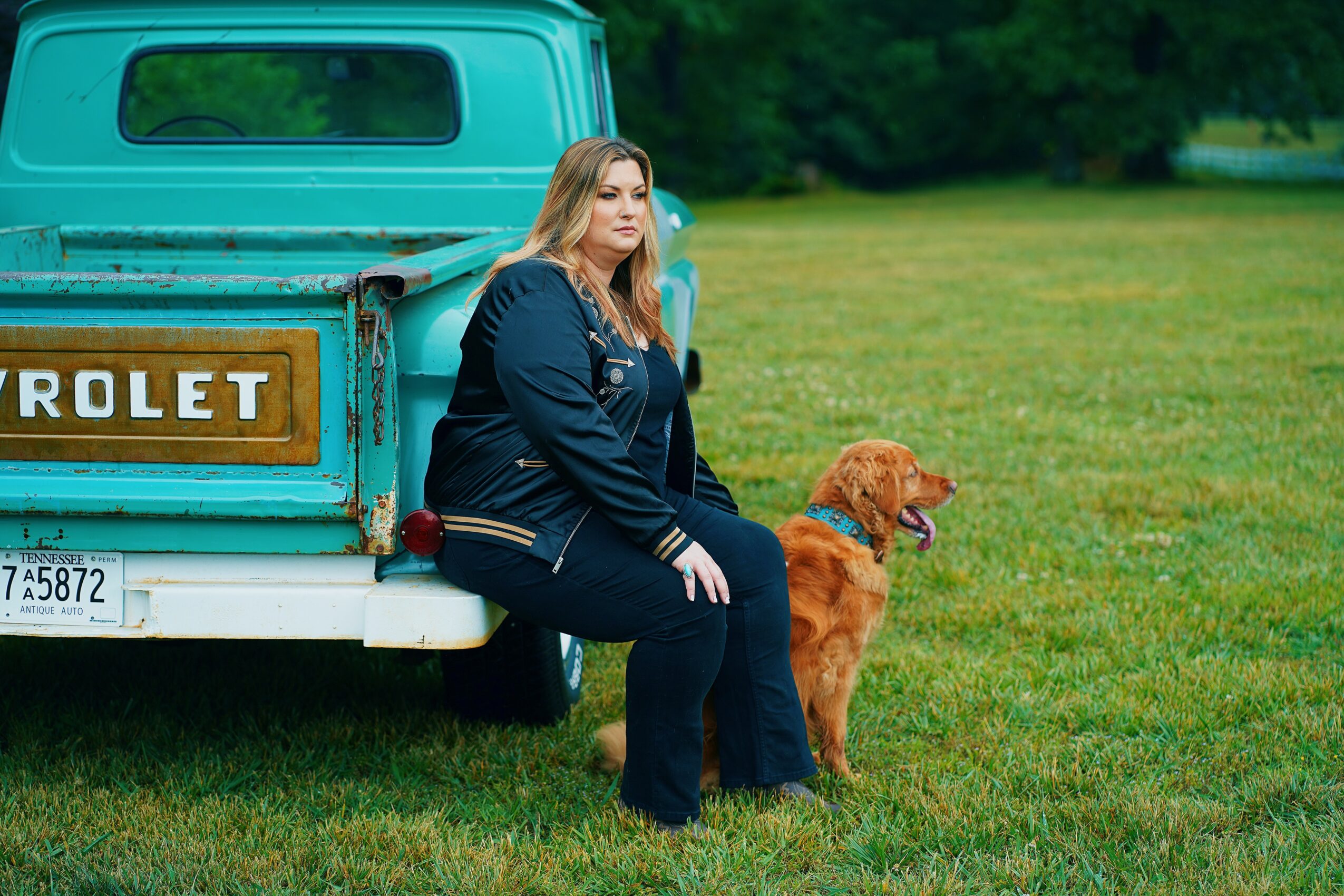 Woman sitting on the back of a vintage Chevrolet truck with a dog on a grassy field.
