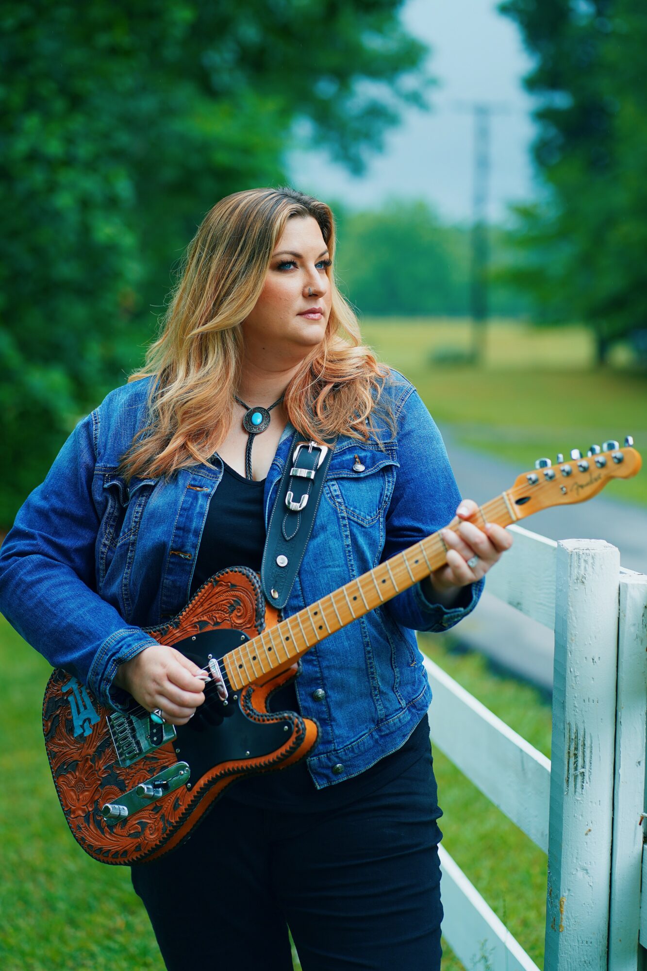 Woman with long hair holding an electric guitar outdoors near a white fence, trees in background.