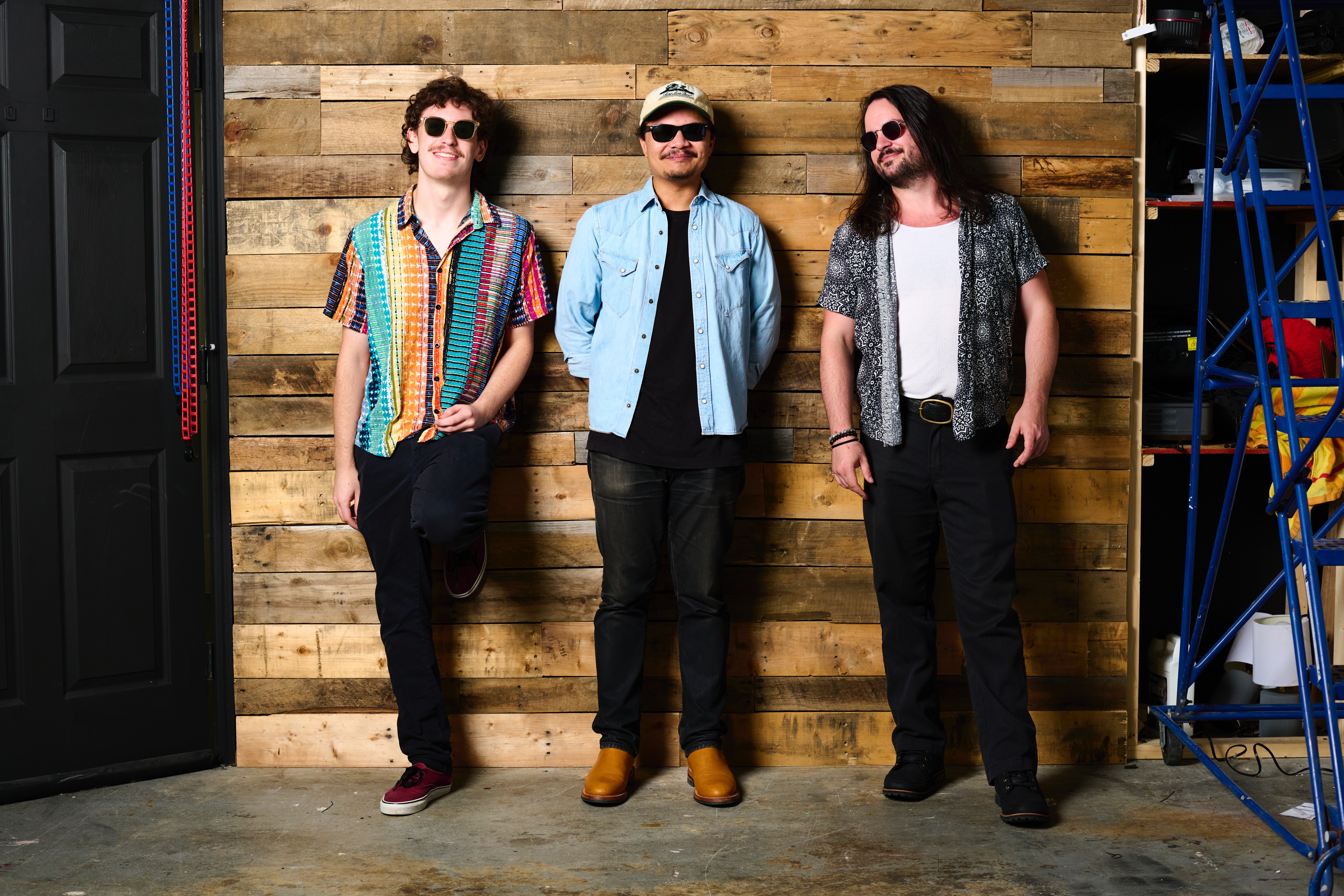 Three people stand against a wooden wall, wearing casual clothing and sunglasses, with a vending machine and blue structure nearby.