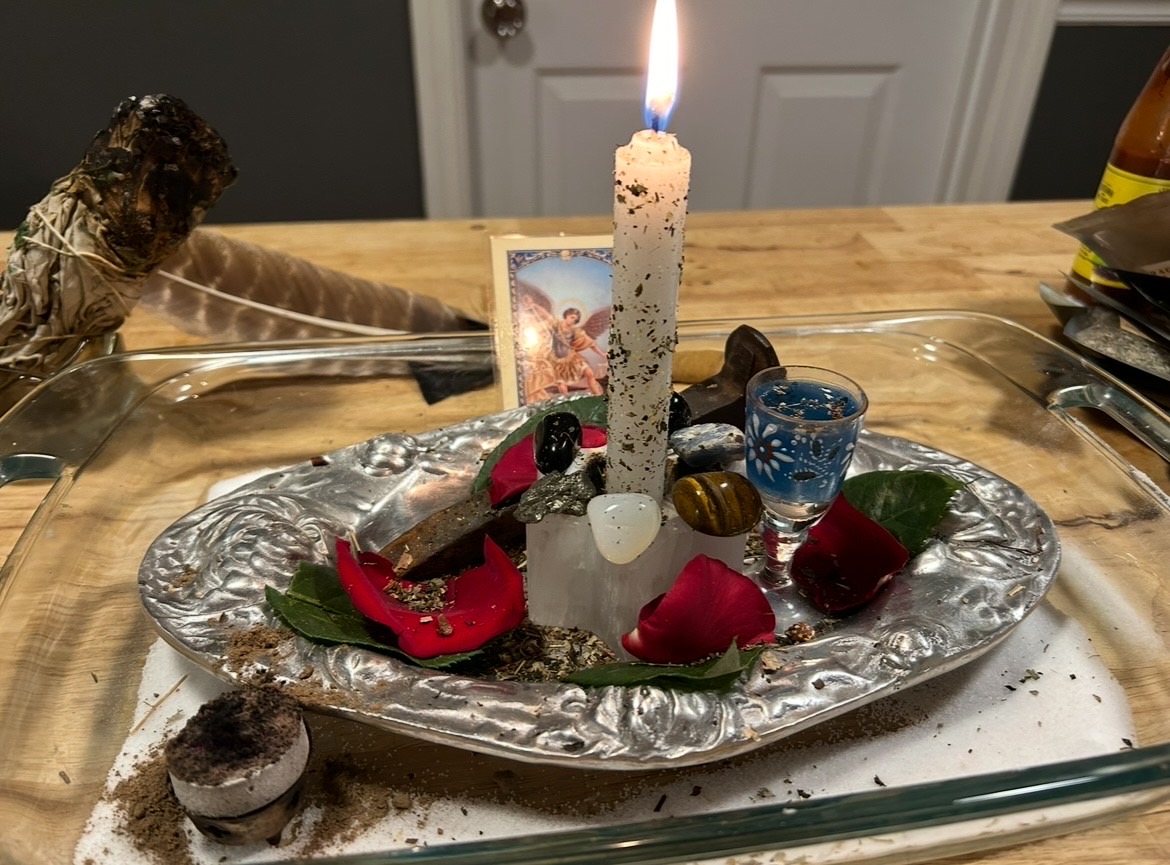 A lit candle on a silver tray with chocolates, rose petals, a small blue glass, and a framed photo, on a wooden table.