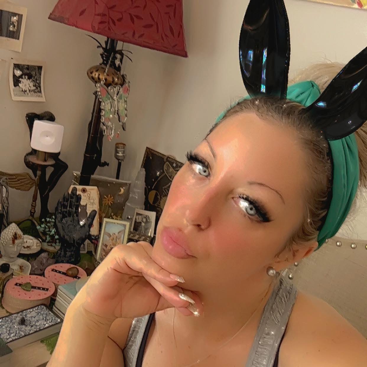 Woman with bunny ears headband, resting chin on hand, in a room with various decorative items and photos