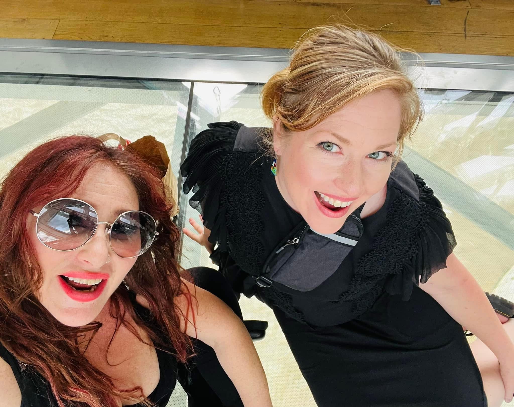 Two women smiling and looking up at the camera, one with sunglasses and red hair, the other with blonde hair, indoors.