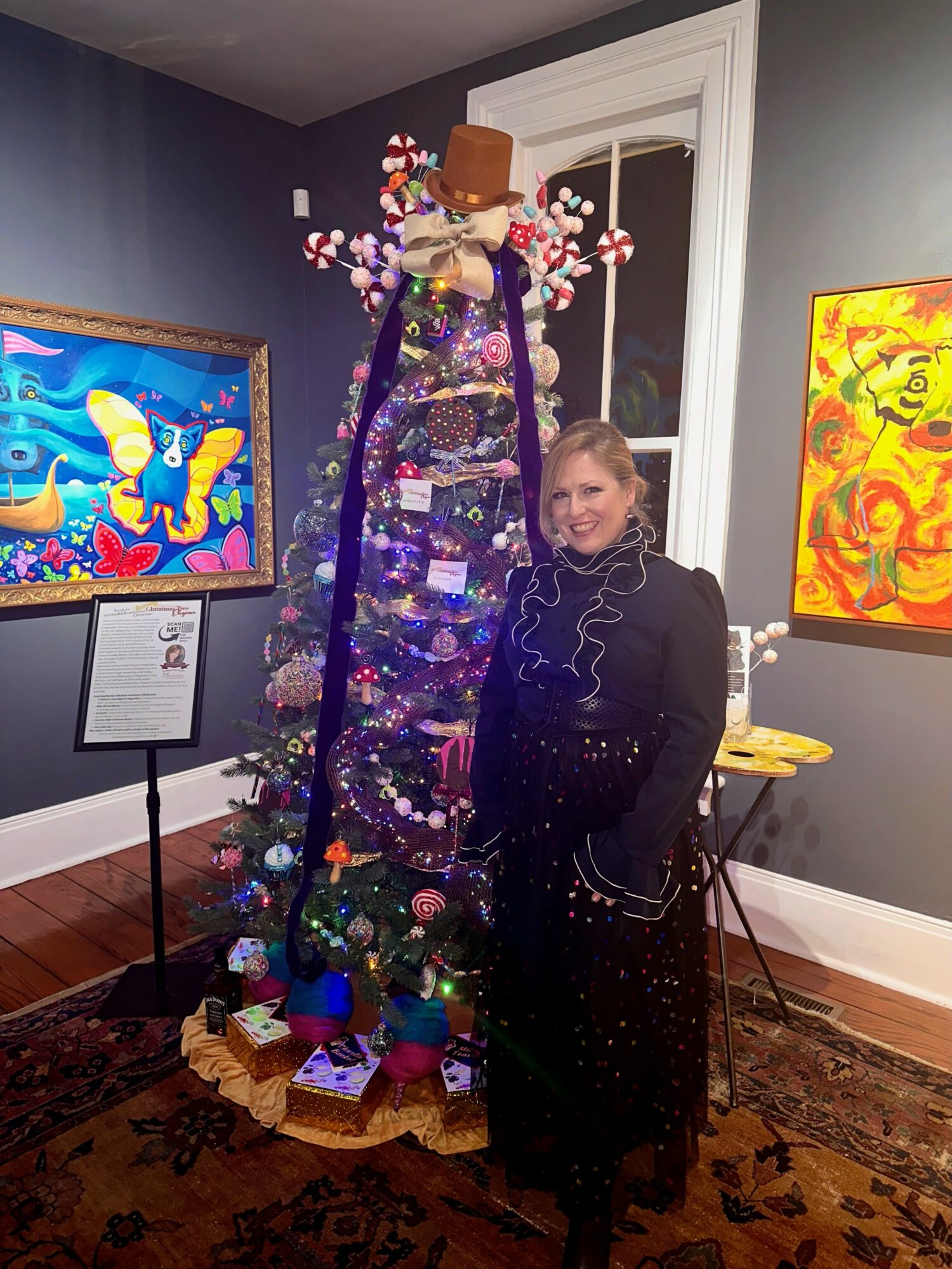 Woman standing next to a decorated Christmas tree in an art gallery, with colorful paintings on the walls.