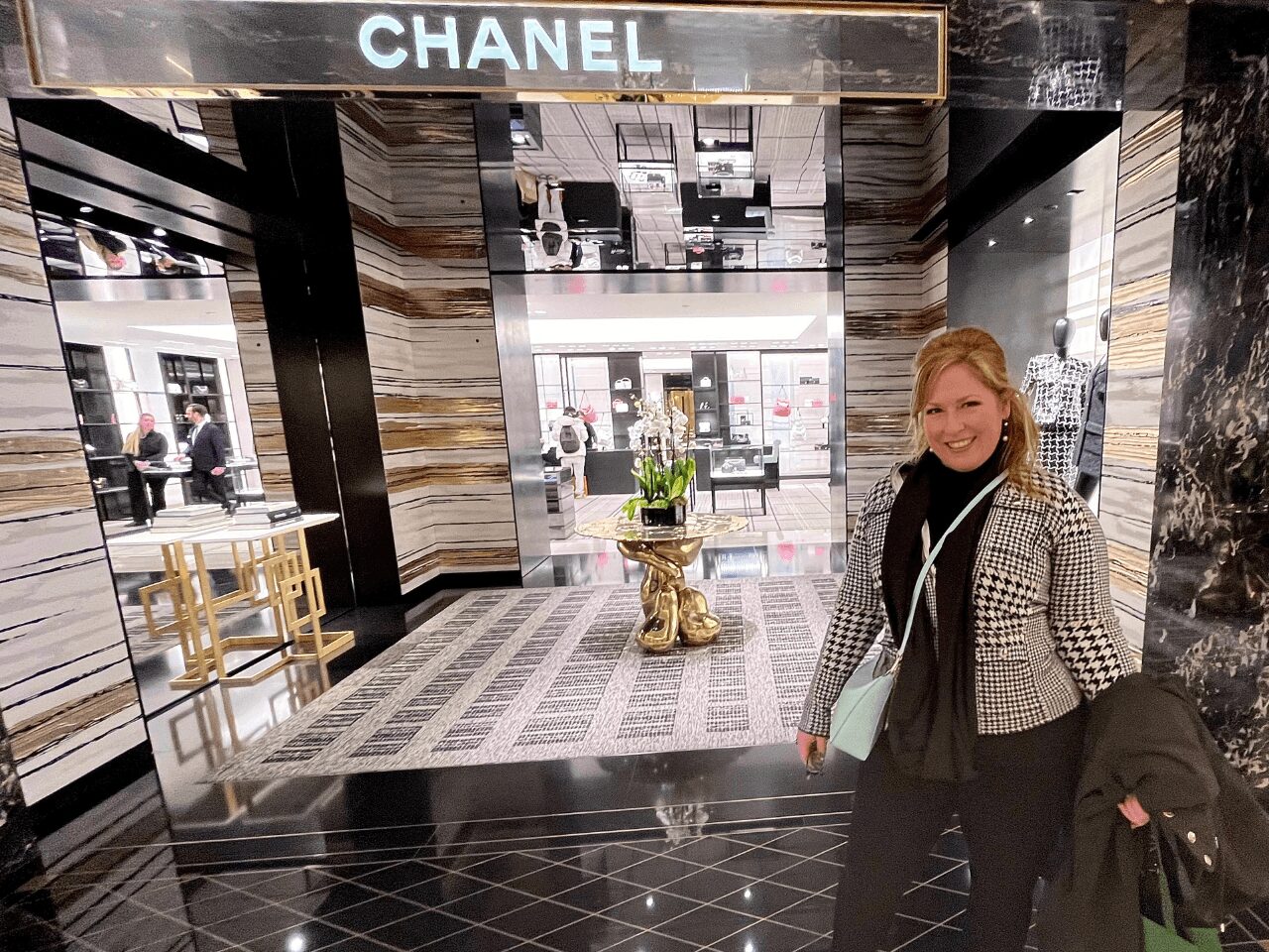A woman with blonde hair smiling stands inside a Chanel store entrance with a marble interior and a potted plant, holding a coat.
