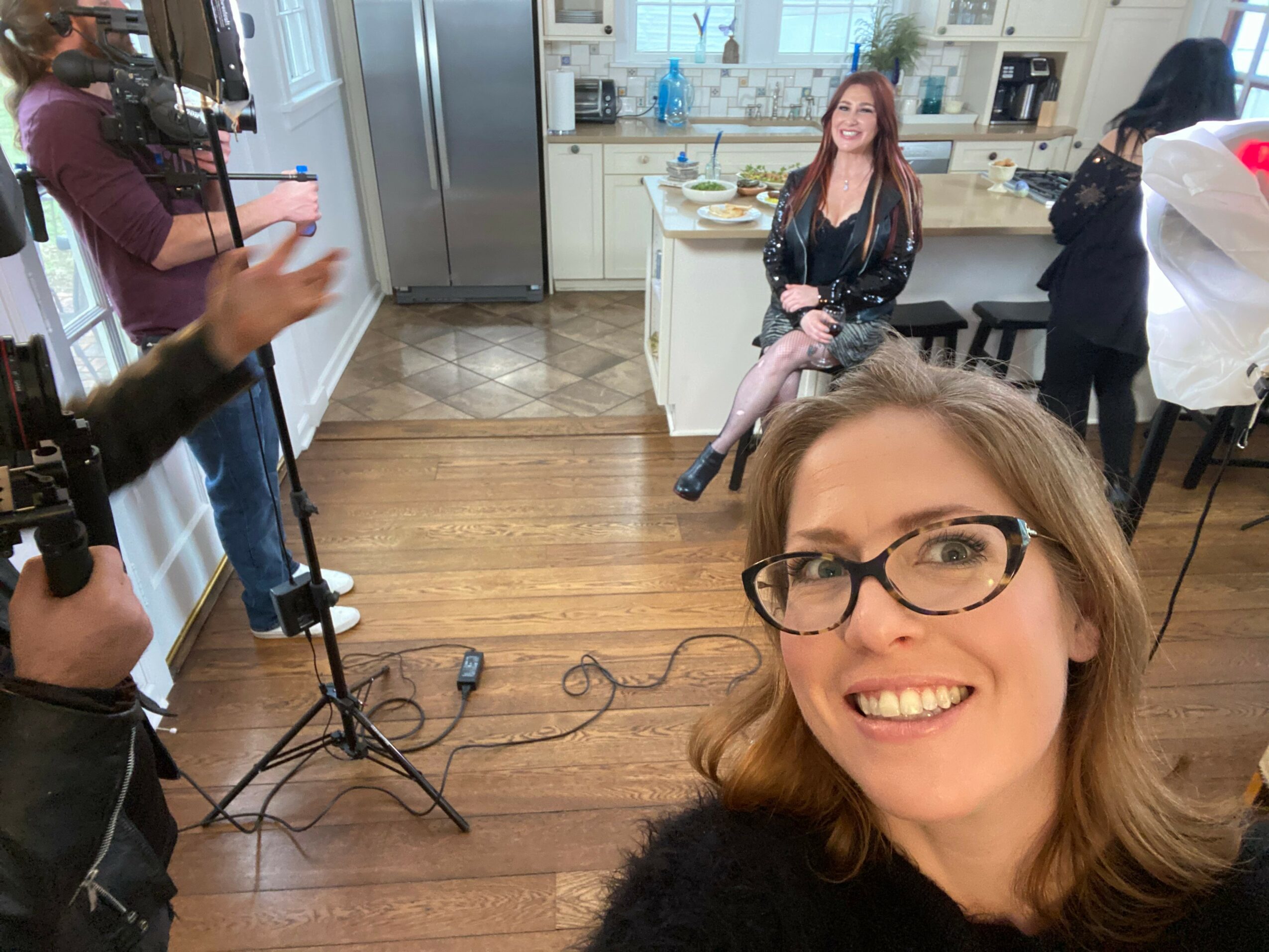 Woman with glasses smiling in a kitchen, taking a selfie with a woman sitting on a counter in the background.