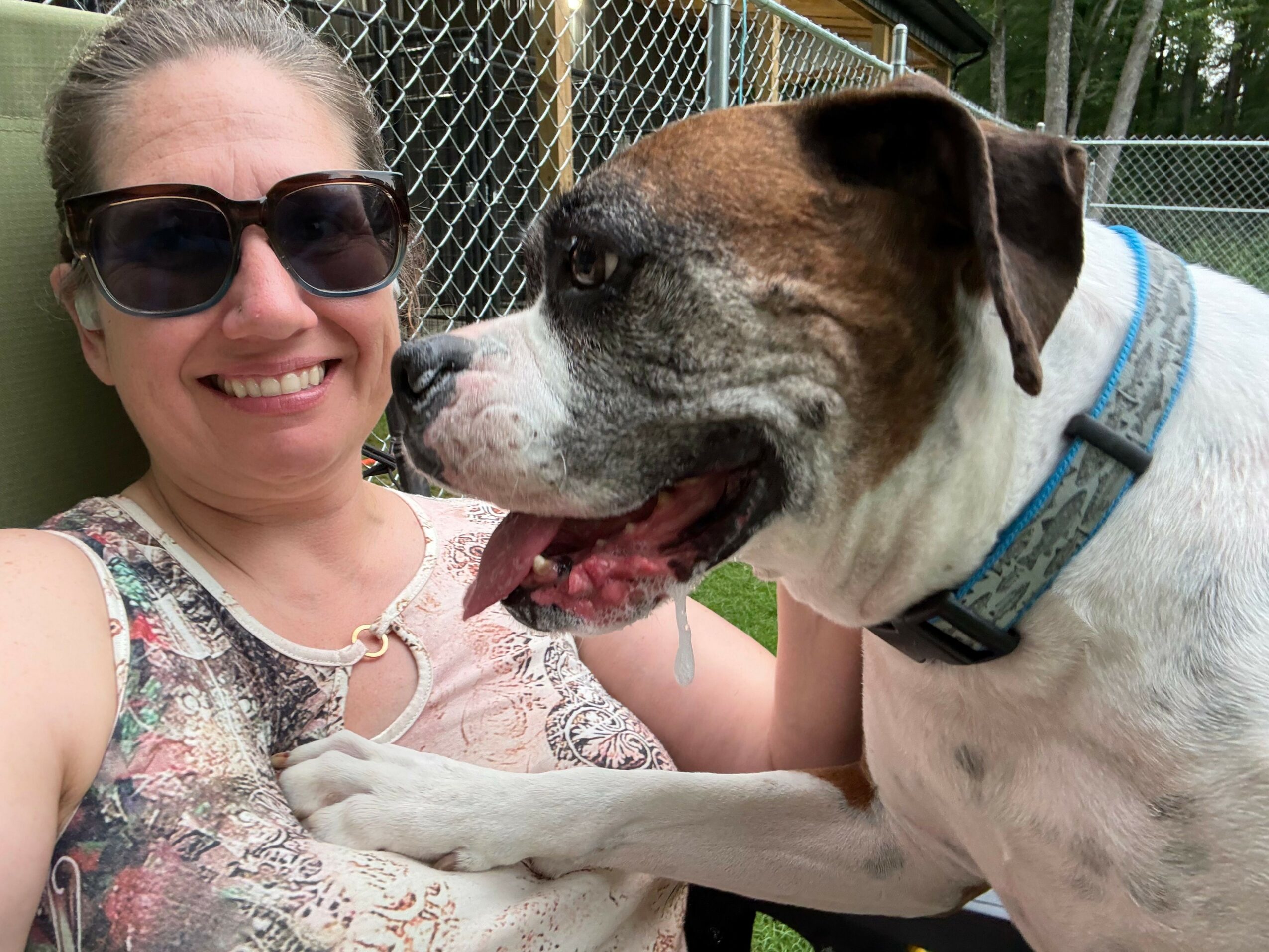 Person lying on grass with a dog, both smiling, person wearing sunglasses and floral top, dog with collar, fence in background.
