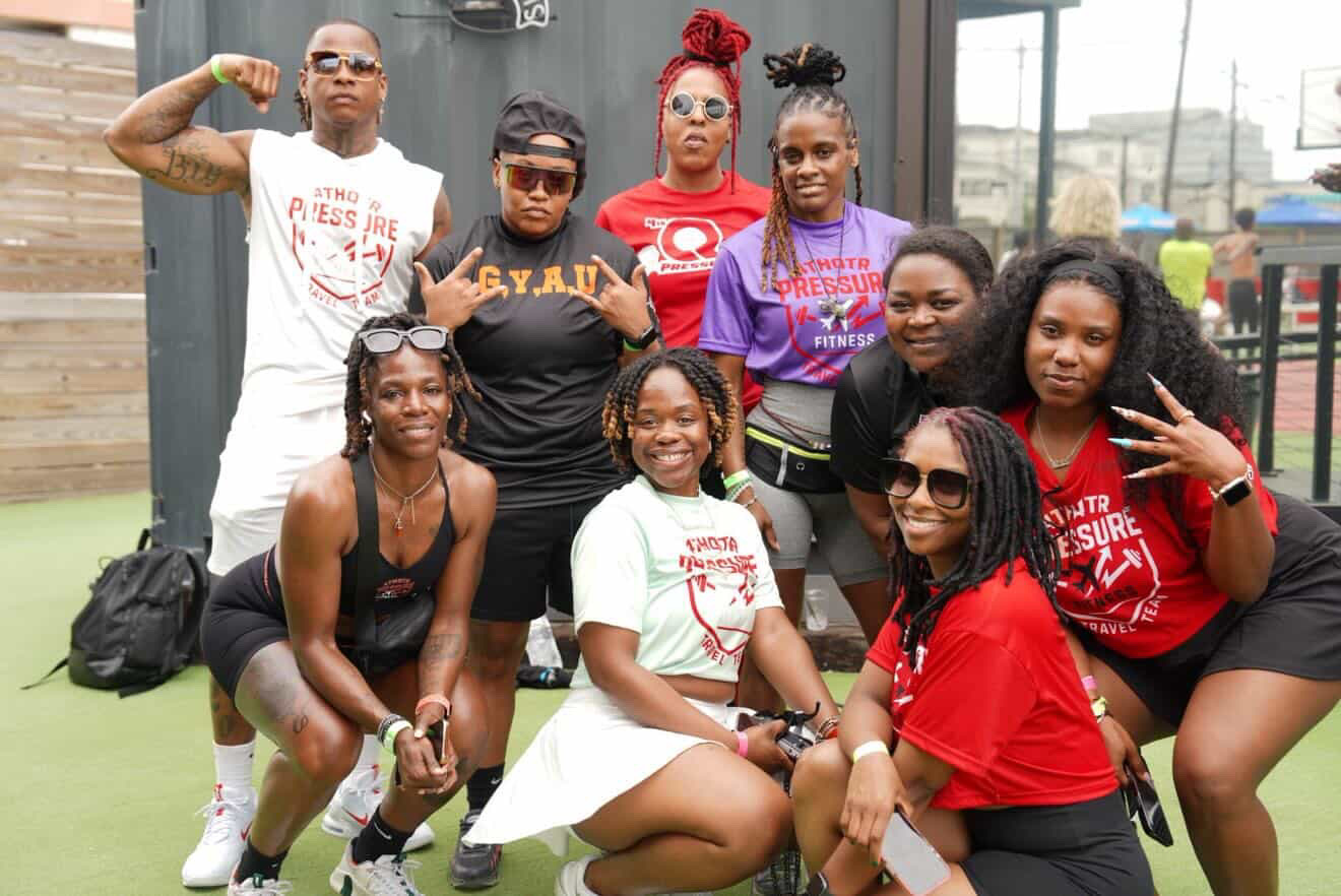Group of nine women posing outdoors, some making hand gestures, smiling, and wearing casual clothing and sunglasses.