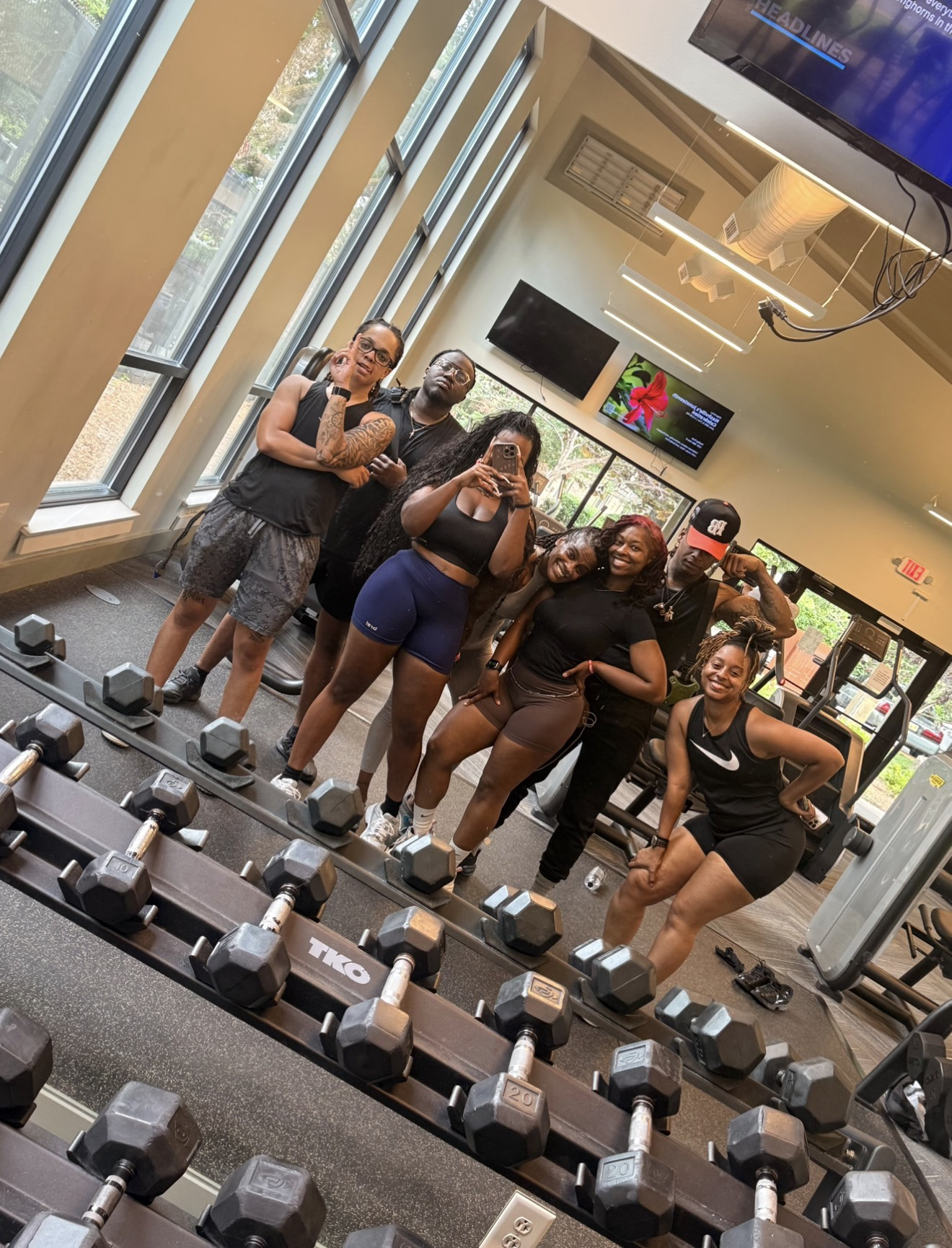 Group of six women taking a selfie in a gym with dumbbells in the foreground, large windows, and screens on the wall.