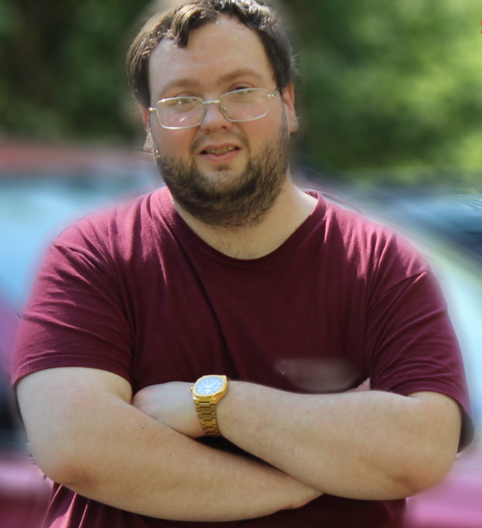 Man with glasses and beard wearing a maroon shirt, arms crossed outdoors with blurred background