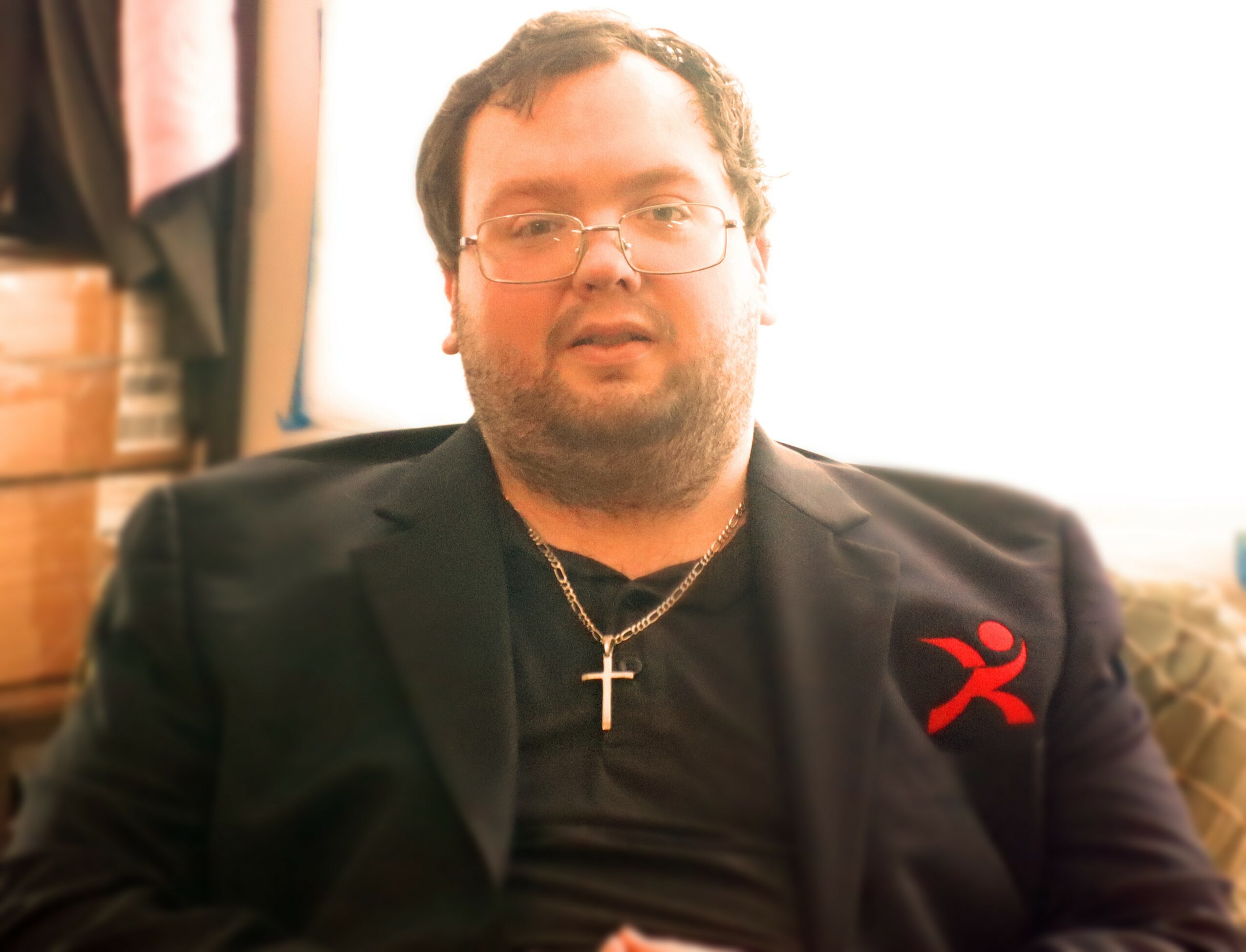 Man with glasses, beard, wearing a black blazer and a cross necklace, sitting indoors with bright light behind him.
