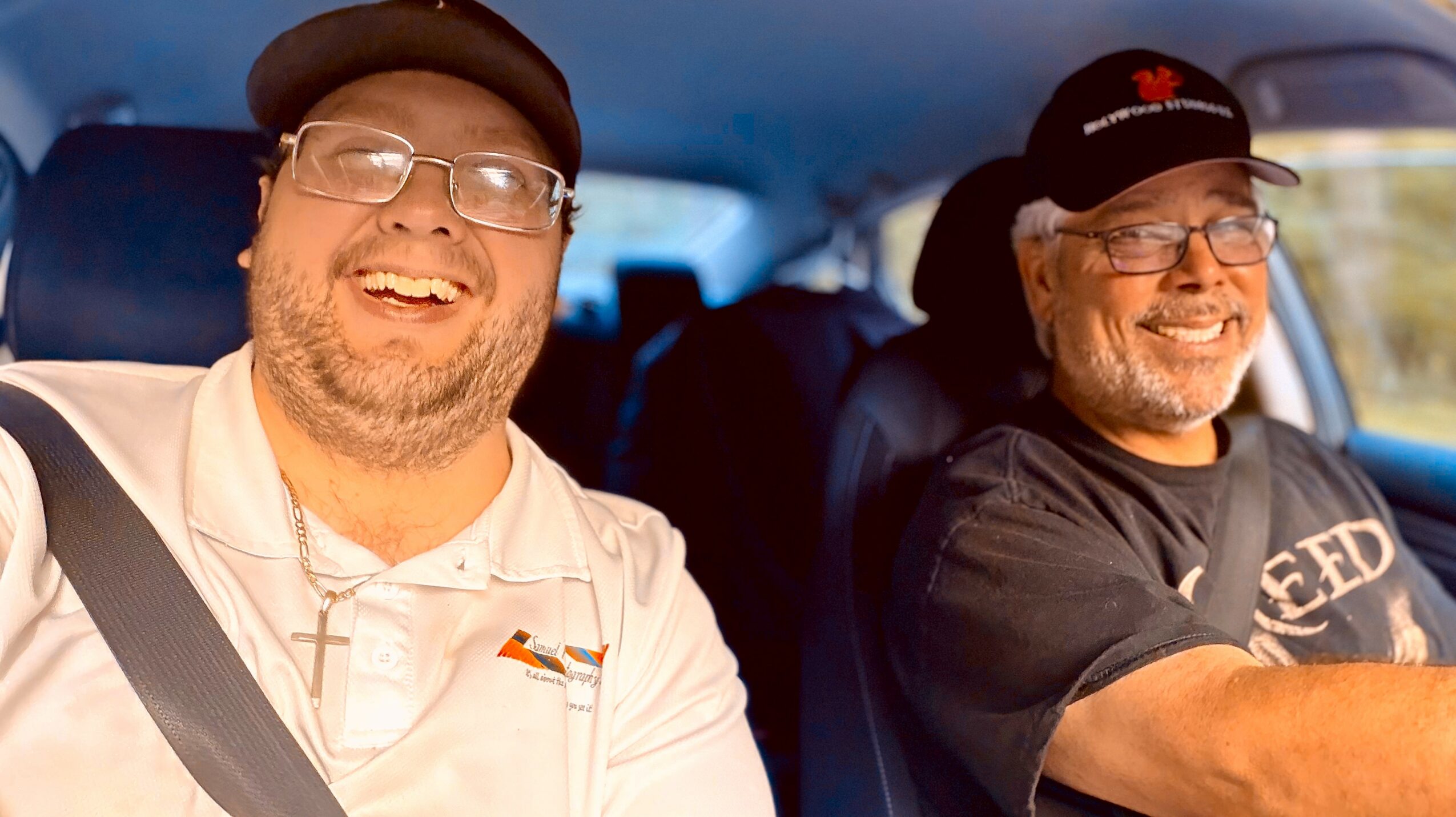 Two smiling men wearing glasses and hats sit in a car, one in the front passenger seat and the other in the driver seat.