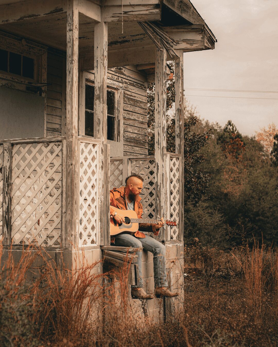 Person sitting on porch of an old, weathered house playing guitar outdoors, surrounded by tall grass and trees.