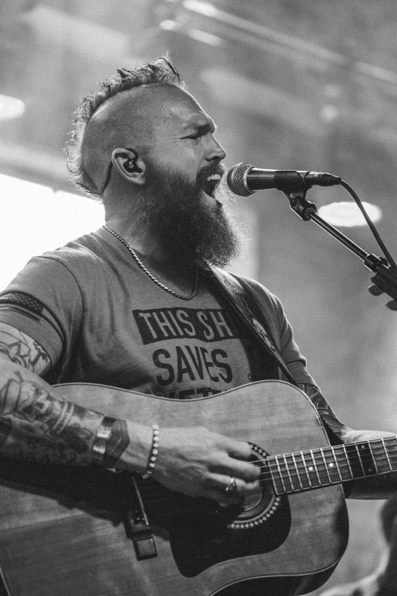 Man with a beard and tattoos singing into a microphone while playing an acoustic guitar, in black and white.