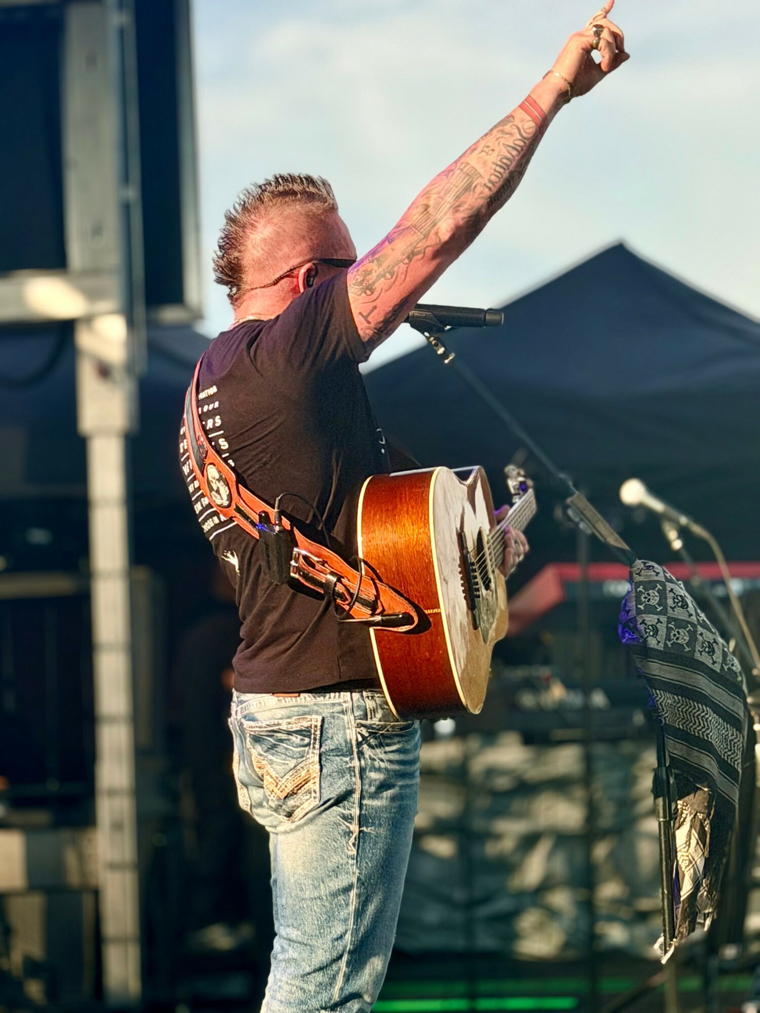 Man with tattoos raising his right arm, playing an acoustic guitar outdoors during daytime.