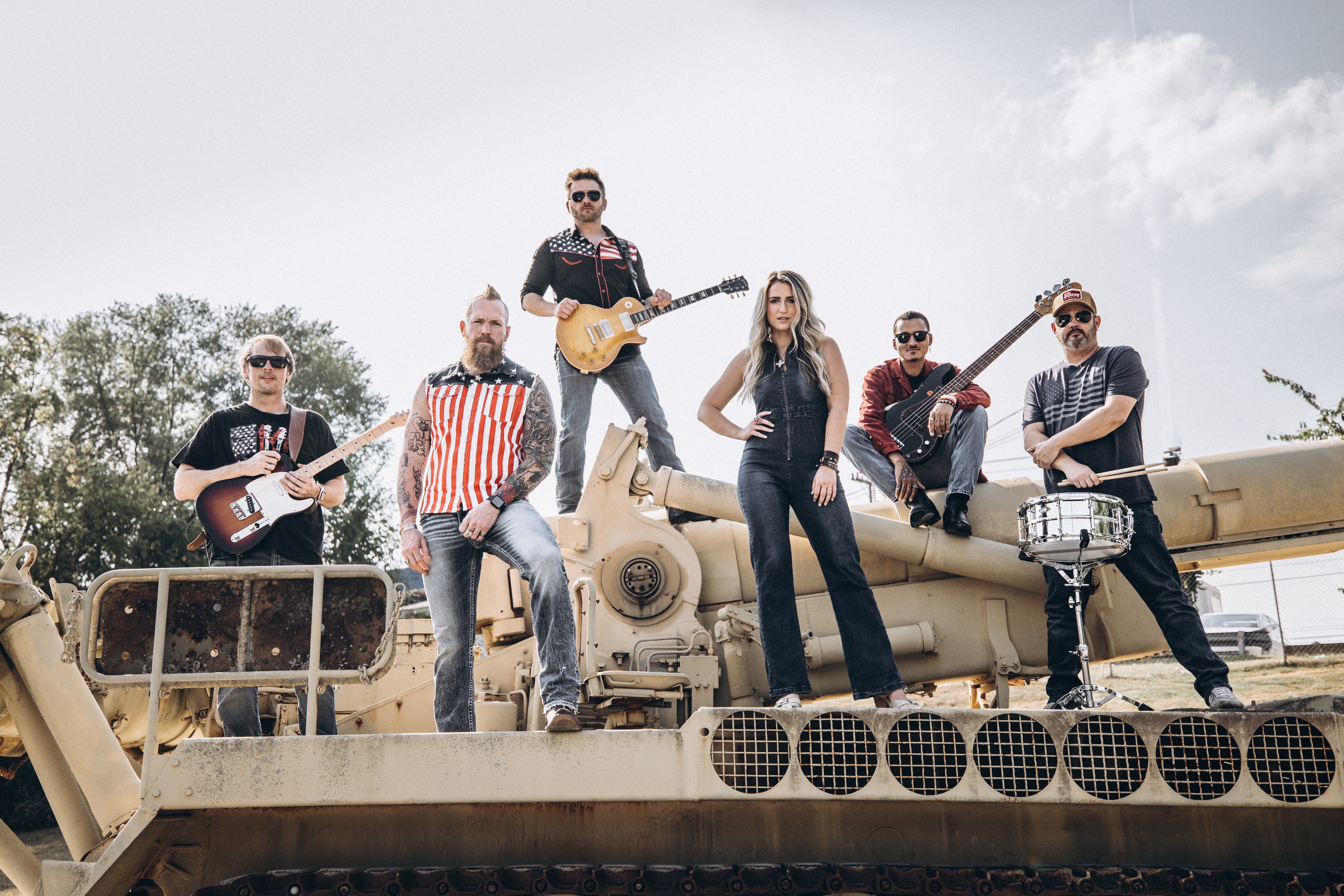 Group of six people standing on a military tank, some holding guitars, outdoors with a cloudy sky.