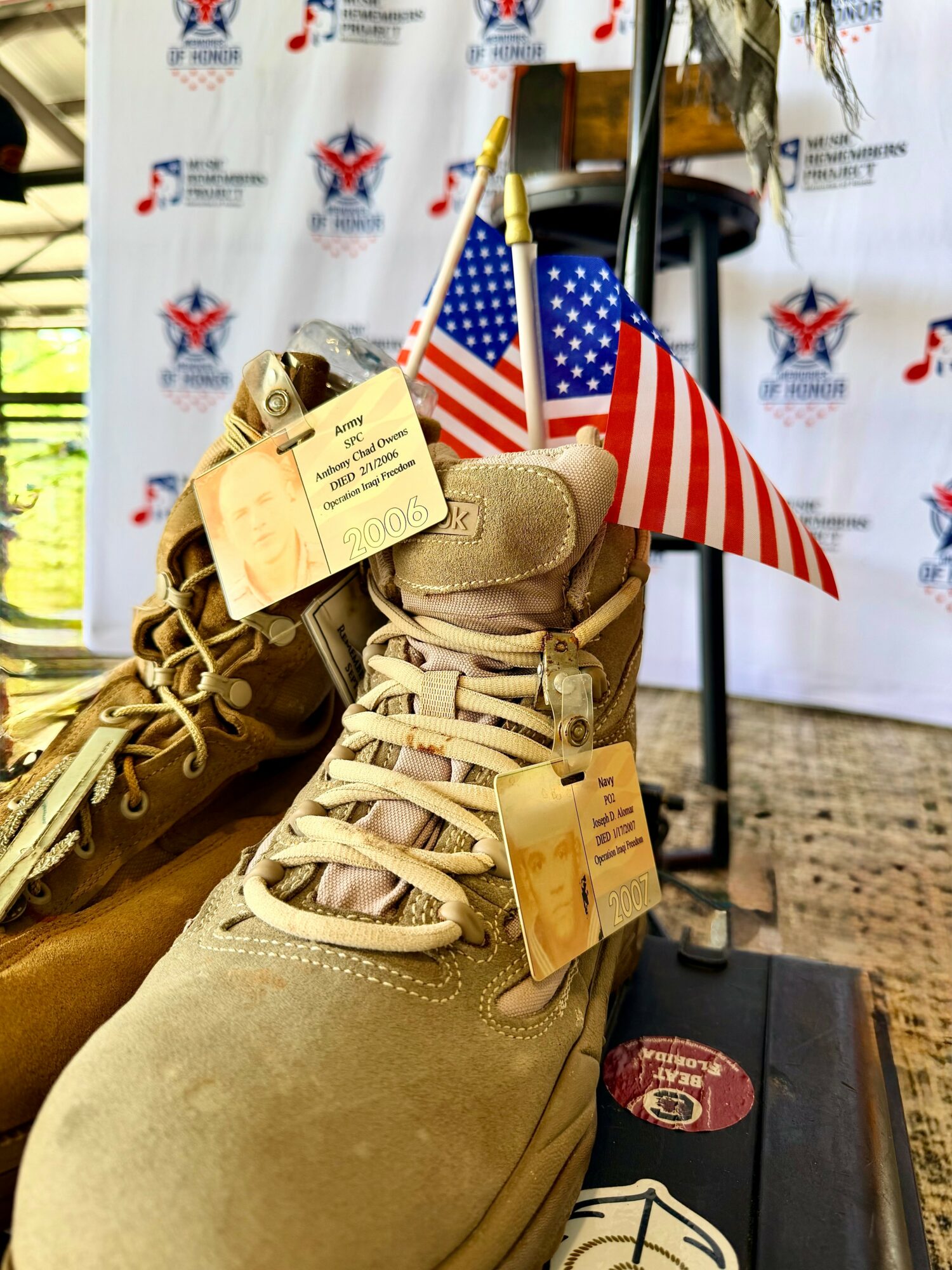 Pair of tan combat boots with tags, American flags in background, and a backdrop with patriotic logos.