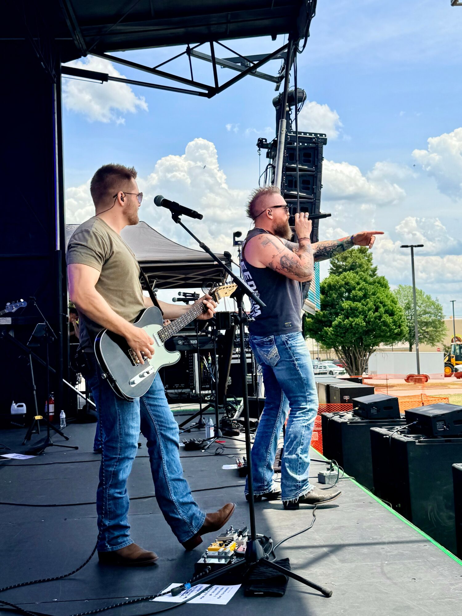 Two musicians perform on an outdoor stage, one singing into a microphone and the other playing guitar, with a blue sky background.
