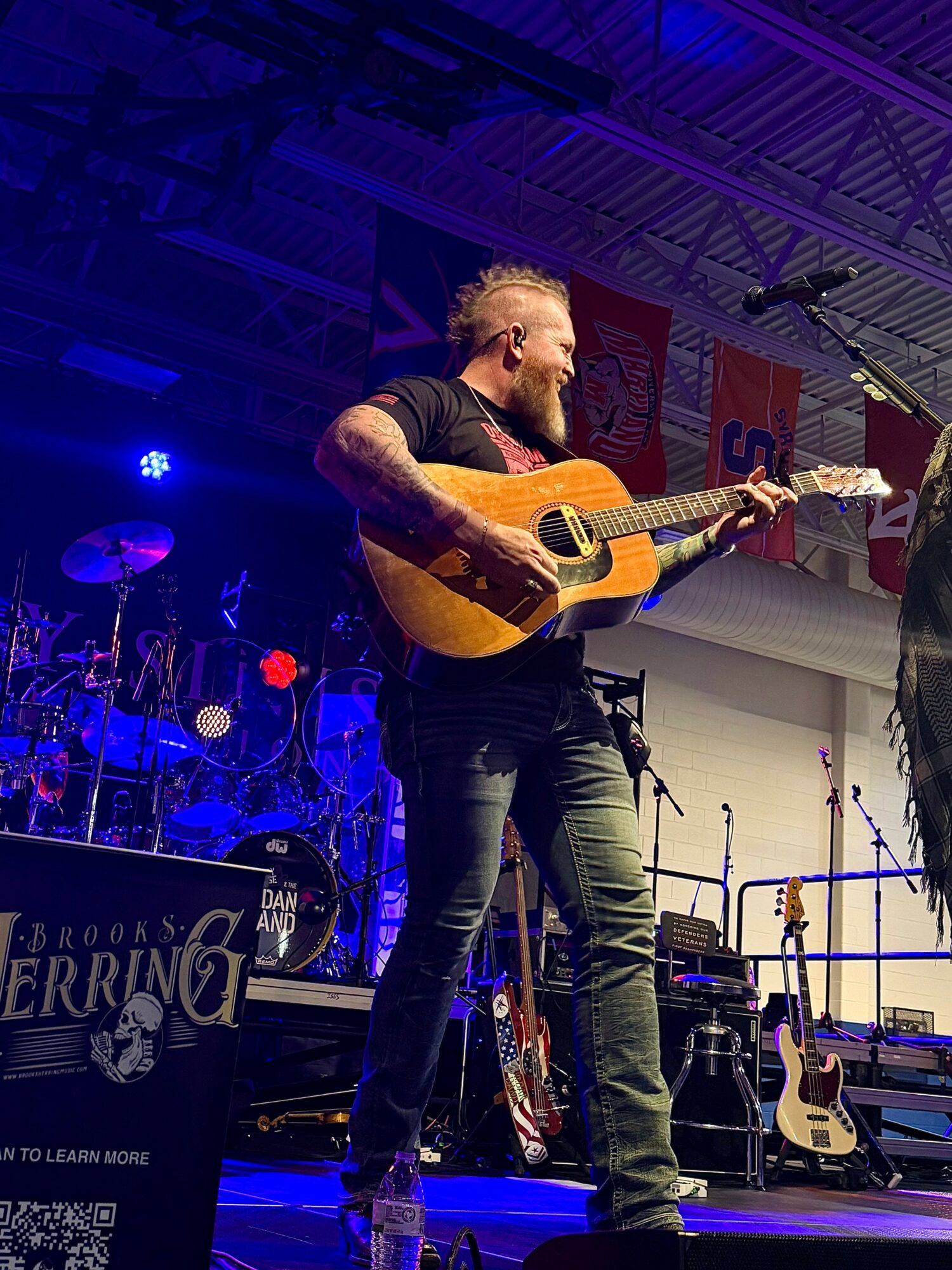 Man with a beard playing an acoustic guitar on stage, with drums and other guitars in the background.