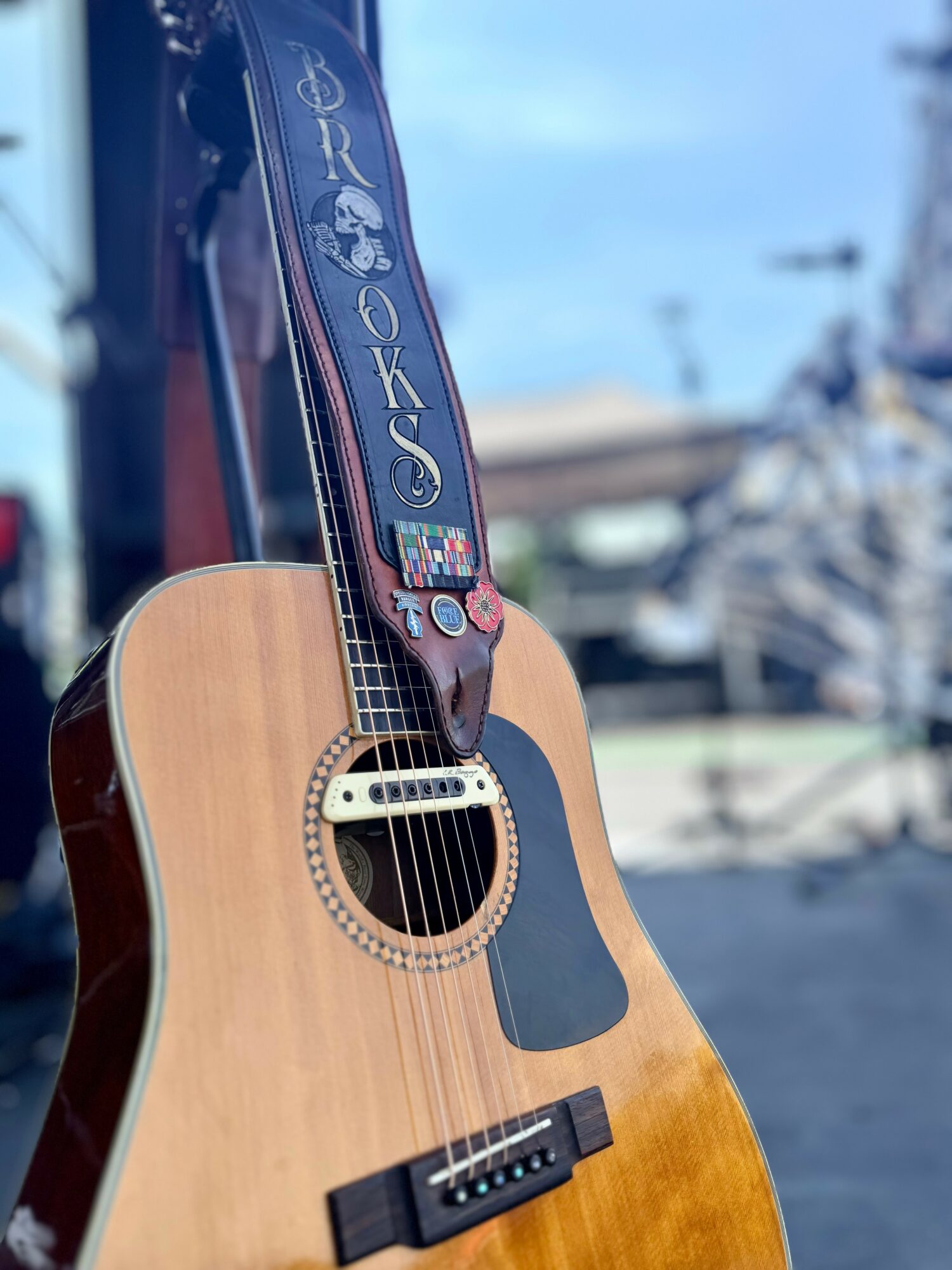Acoustic guitar with a strap hanging over it, outdoors on a sunny day, background shows a street scene.