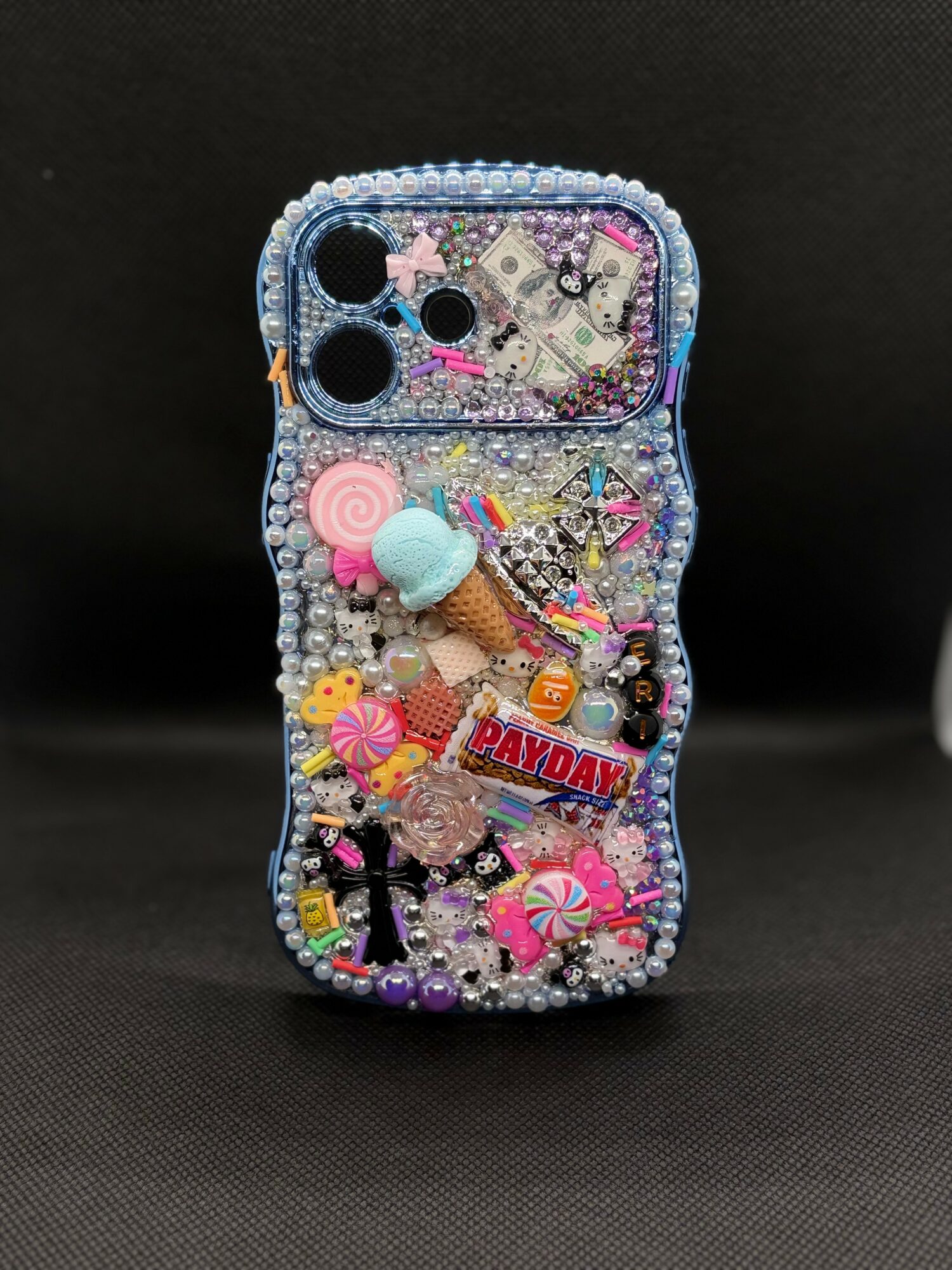 Decorated phone case with beads, charms, and small toys, including ice cream, lollipop, and birthday-themed items, on a black background.