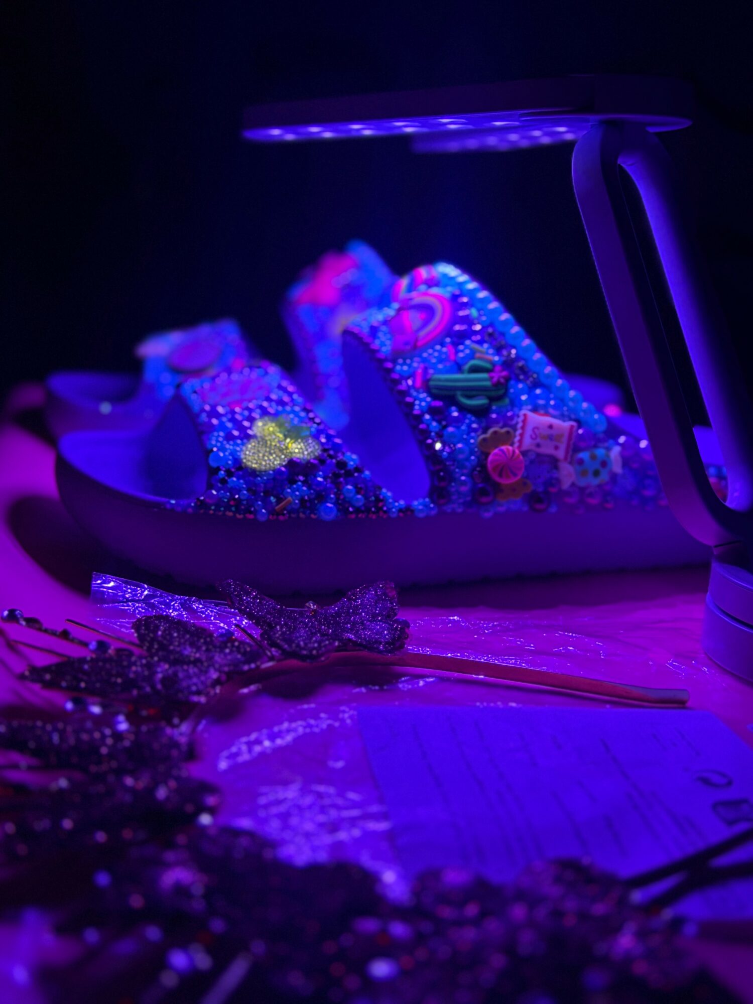 Shoes decorated with colorful beads and accessories under purple lighting, with jewelry and a table edge visible, in a dark setting.