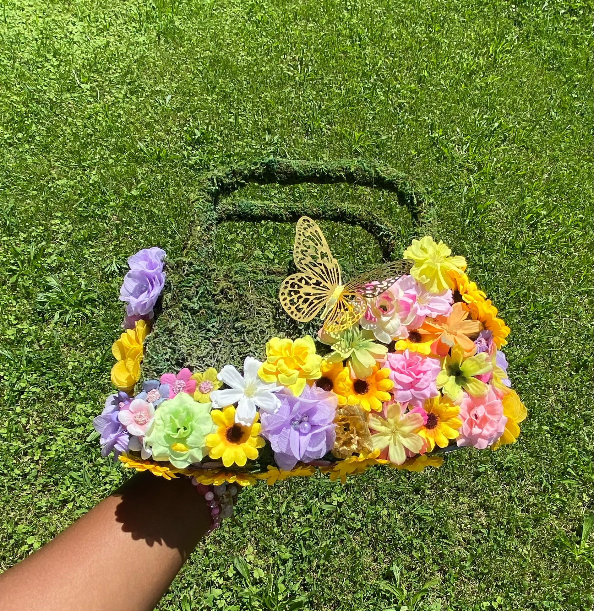 Hand holding a floral handbag with a butterfly decoration, set against a grassy background.
