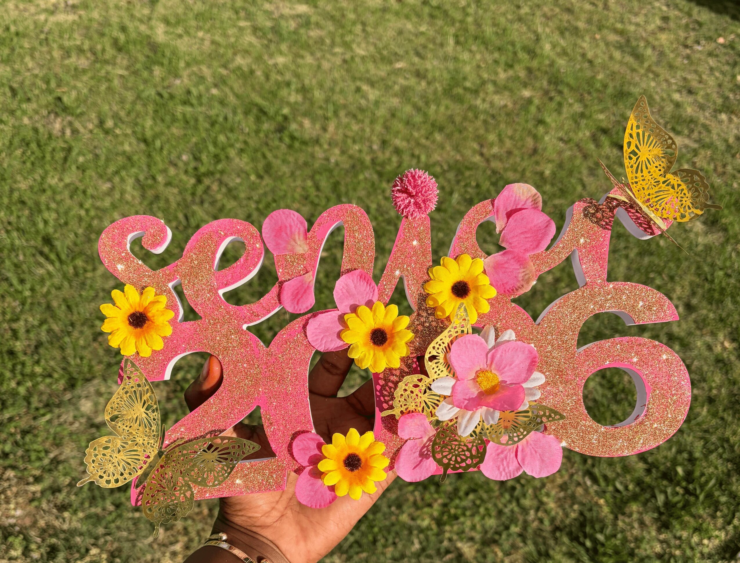 Hand holding pink decorative sign with flowers and butterflies on grass background.