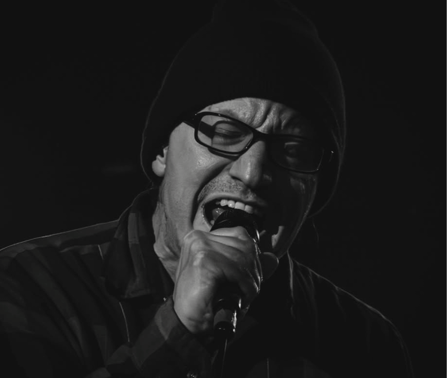 Person singing into a microphone, wearing glasses and a beanie, with an intense facial expression.