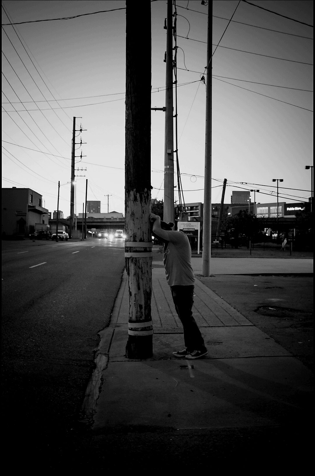 Person leaning against a utility pole on a city sidewalk, with power lines and buildings in the background.