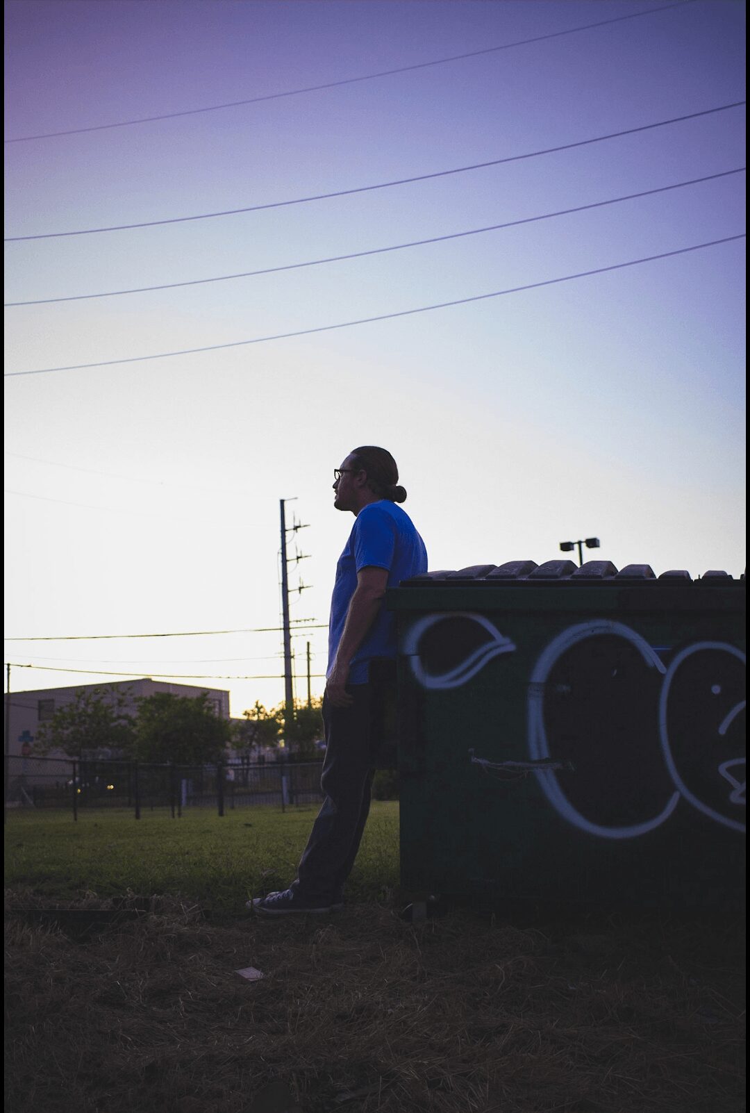 Person standing outdoors near graffiti-covered structure during sunset, with power lines overhead and trees in background.