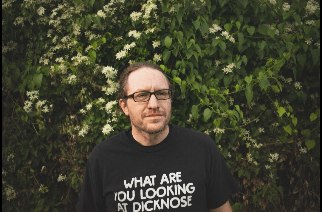 Man with glasses and beard standing outdoors in front of green foliage, wearing a black shirt with white text