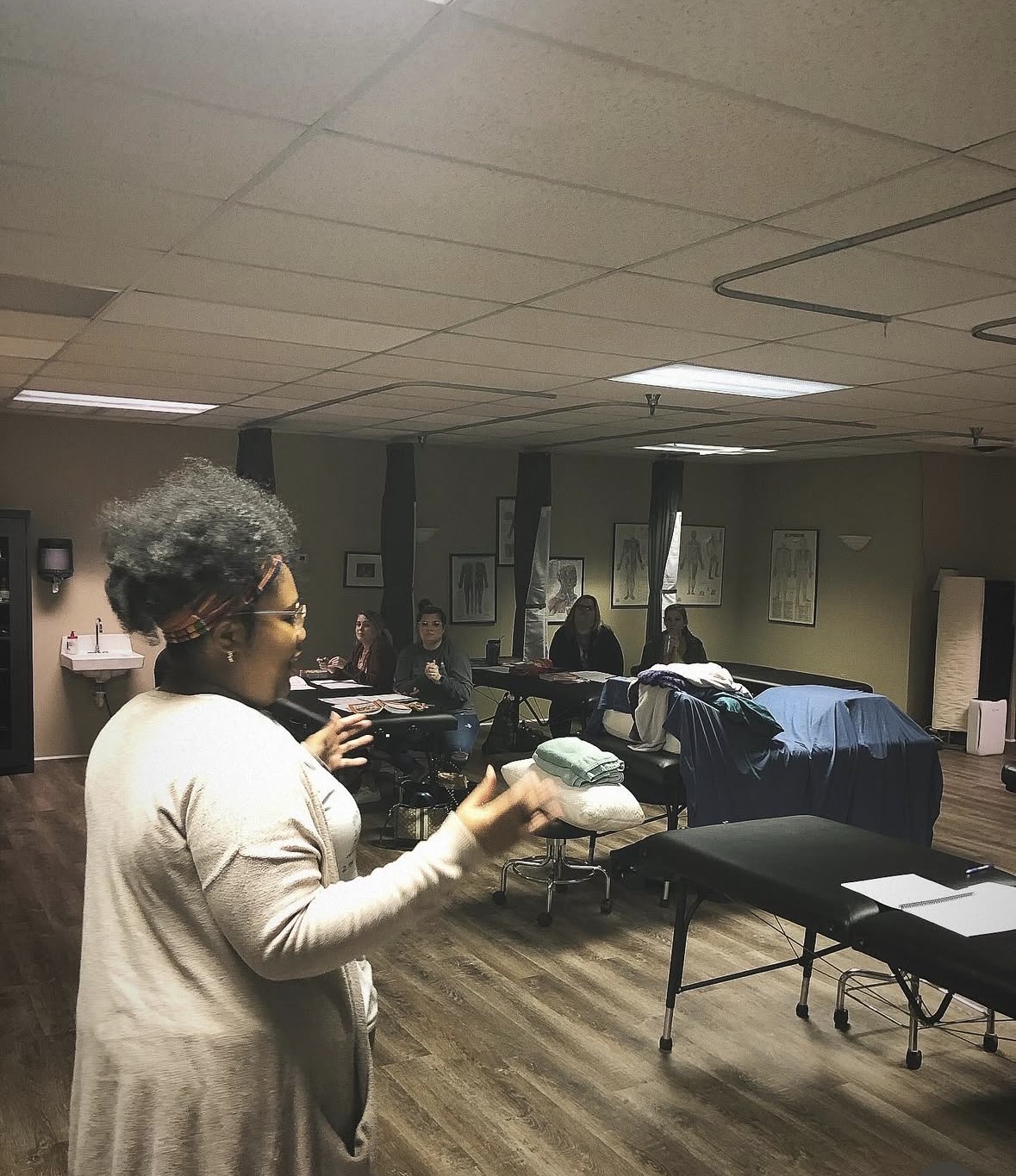 Woman with curly hair and glasses speaking to a group in a room with tables and beds, some people seated in the background.