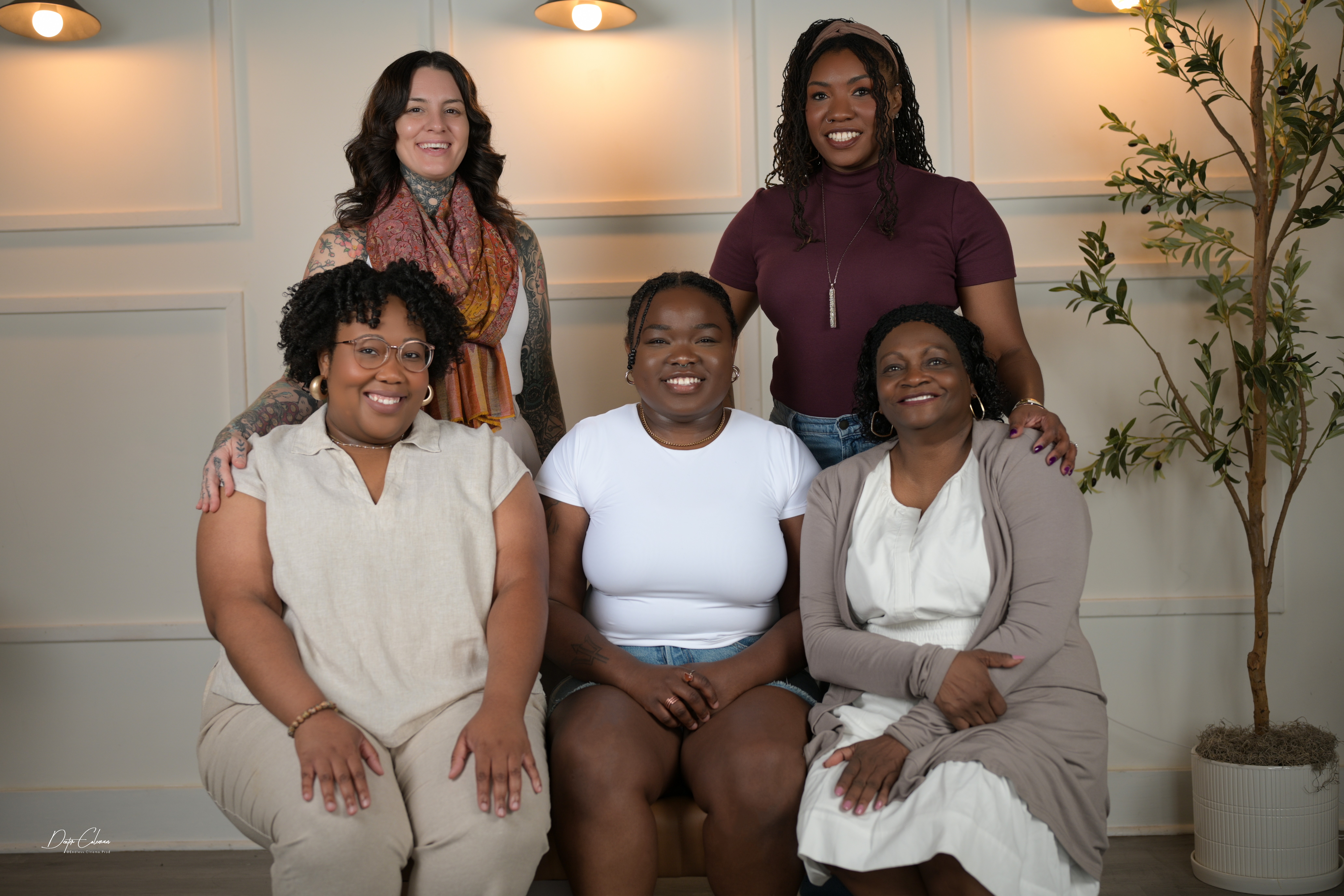 Five women smiling and posing together indoors, with a potted plant and wall decor in the background.