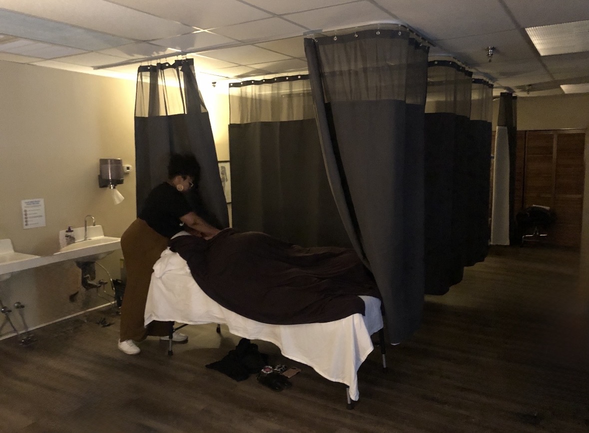 Person giving a massage in a room with black privacy curtains, a bed, and a small table with supplies.