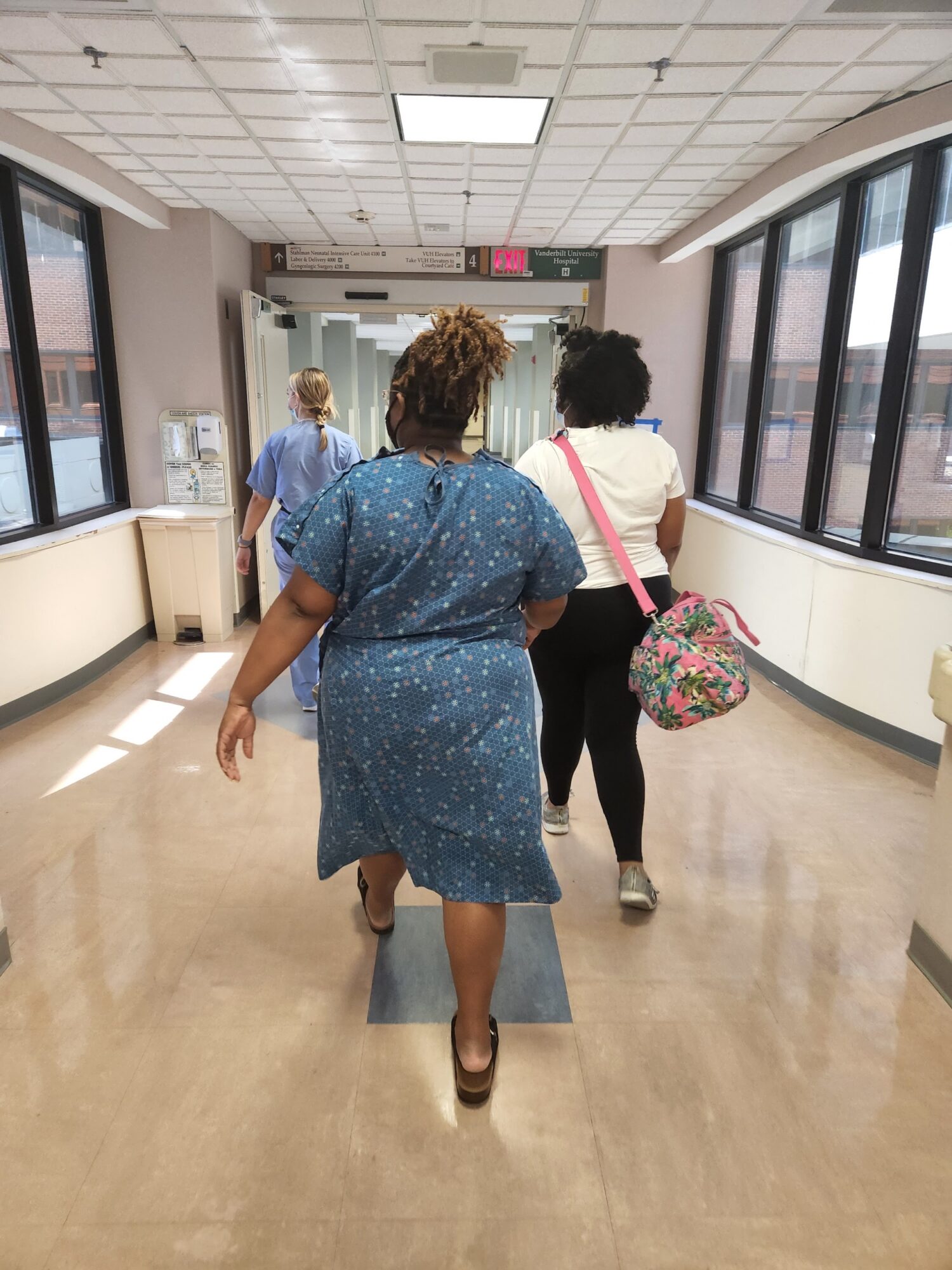 Three women walk through a brightly lit corridor with large windows, one carrying a floral bag, others with curly hair.