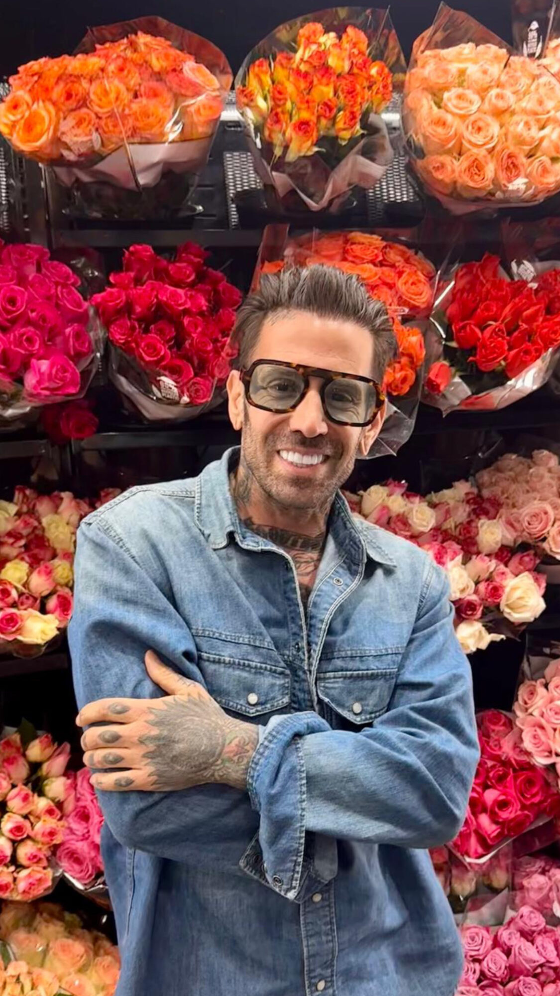 Man with sunglasses smiling in front of flower bouquets, wearing a denim jacket, arms crossed.