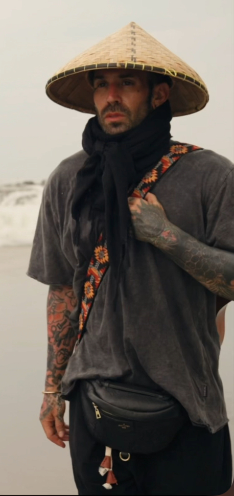 Person wearing a conical hat, dark scarf, and casual clothing, standing outdoors near water with waves in the background.