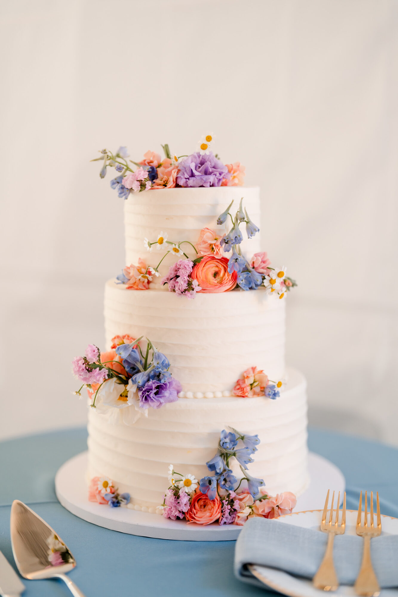 Three-tiered white wedding cake decorated with colorful flowers, on a blue table with gold utensils and a napkin.